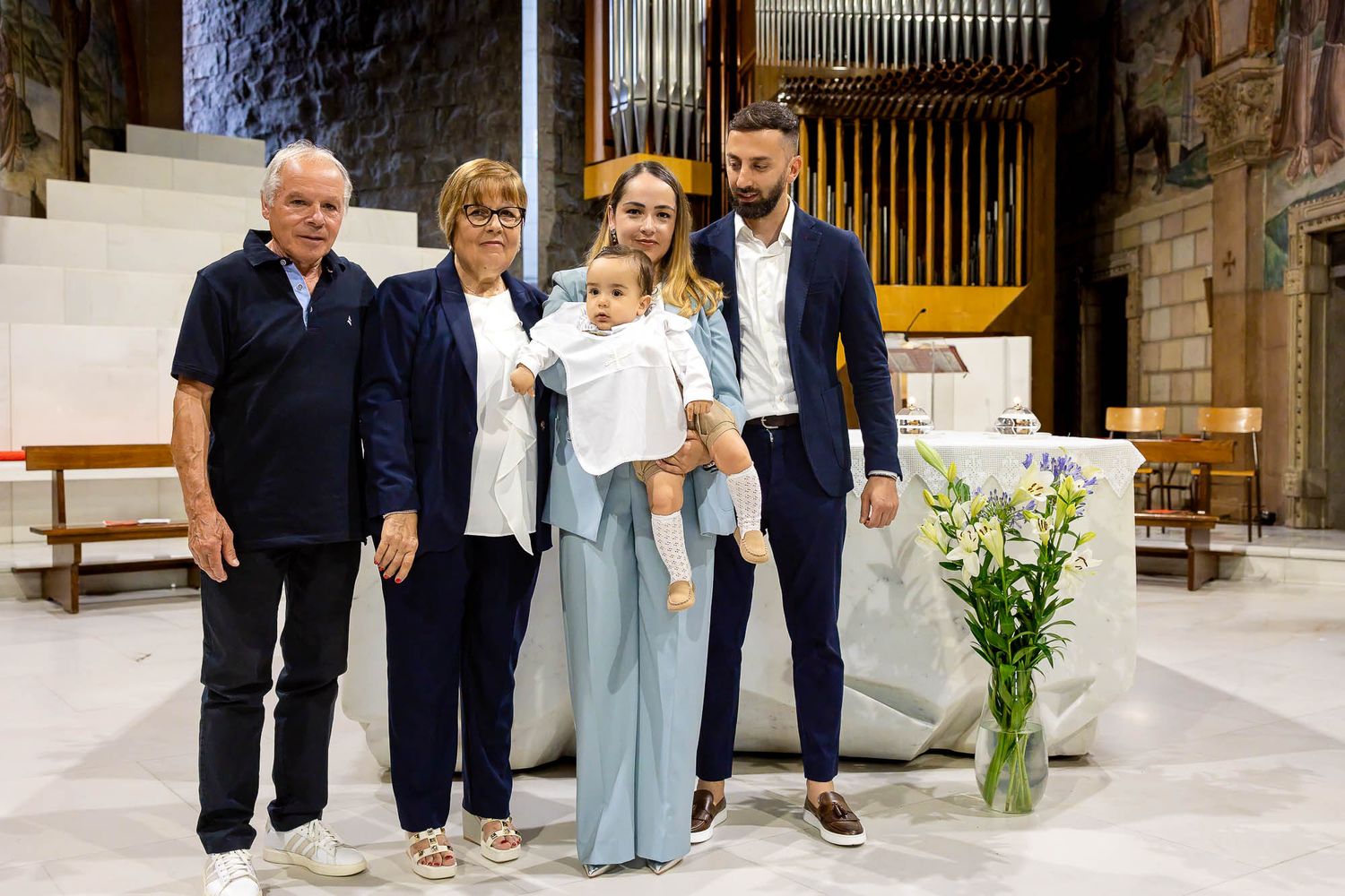 Family posing together in a church during a baptism ceremony, with parents holding a baby dressed in white.