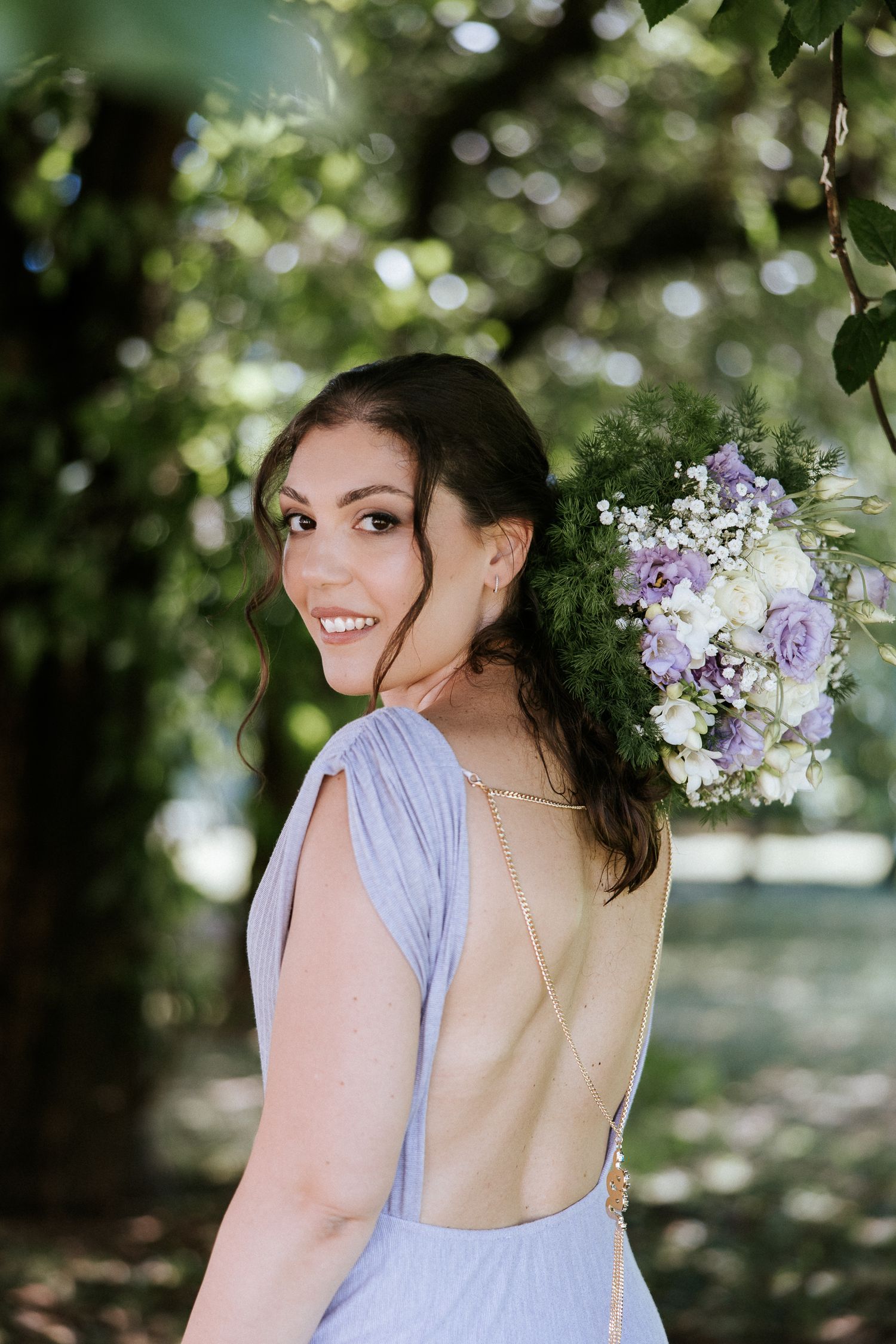 A woman in a lavender dress poses outdoors against a backdrop of blooming lilac flowers and green foliage.