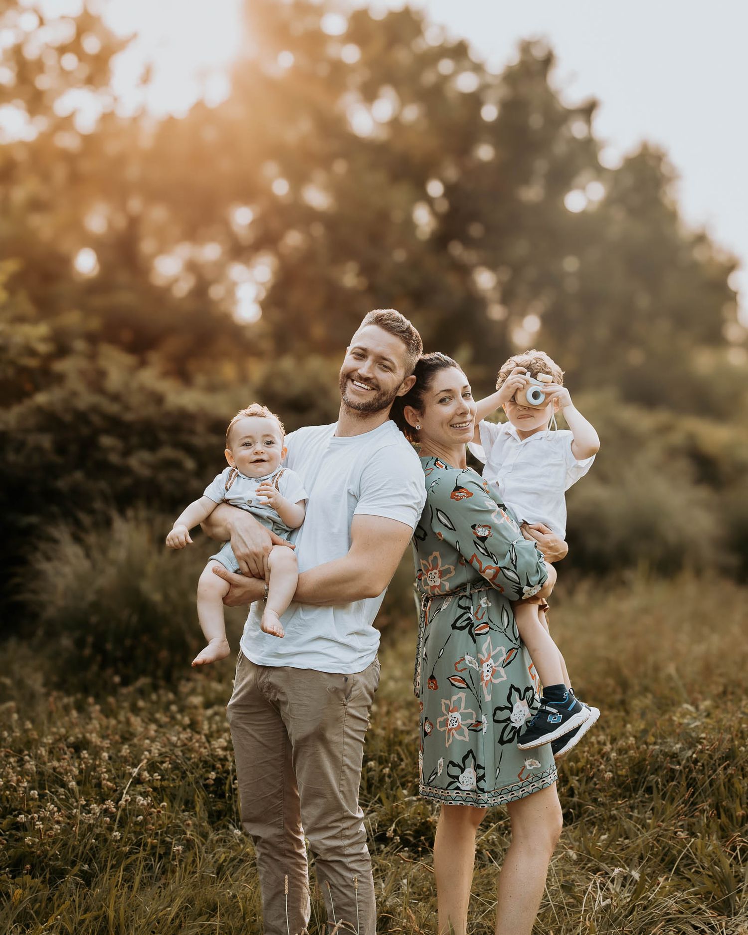 Happy family with two babies posing outdoors in golden hour light, smiling couple holding toddlers in a sunny meadow.