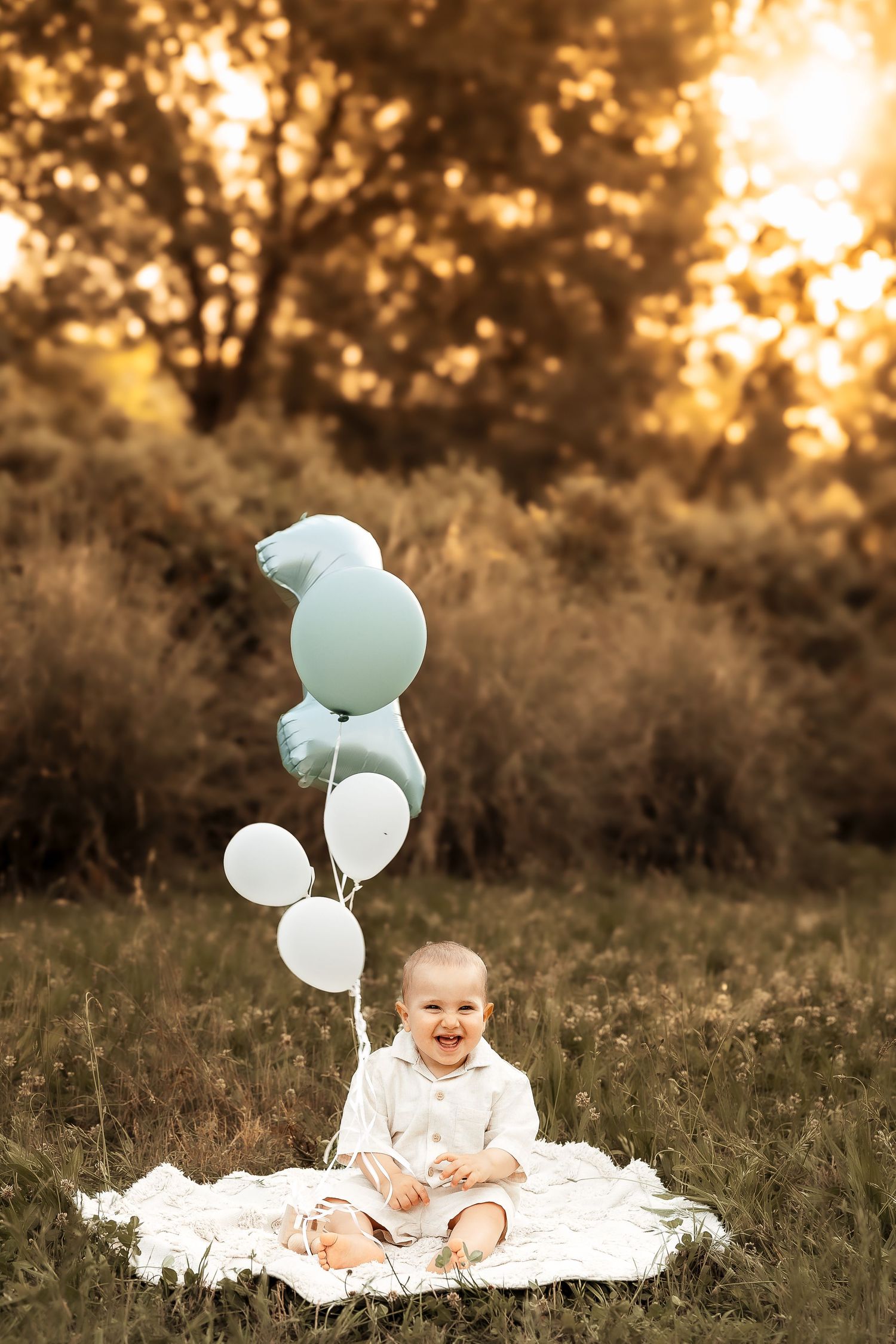 Bambino durante un servizio fotografico per il primo compleanno seduto su una coperta all’aperto circondato da palloncini