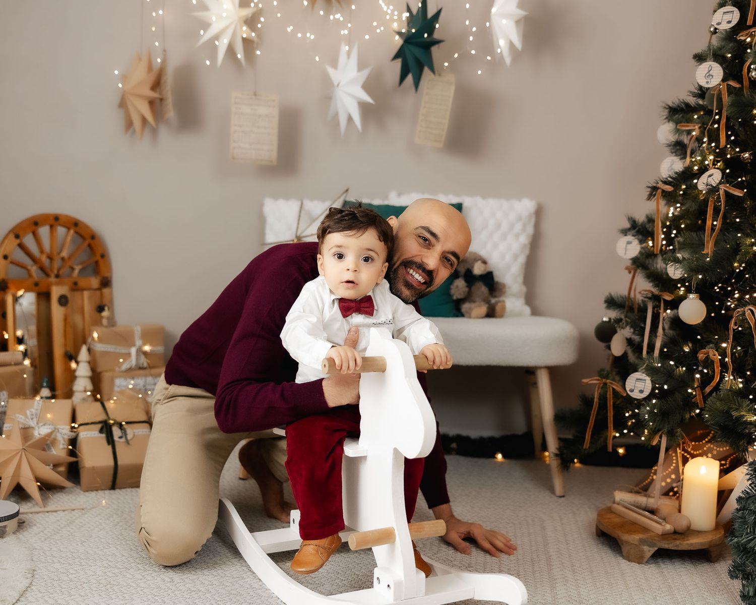 Father and baby boy pose with white rocking horse near Christmas tree and holiday decorations.