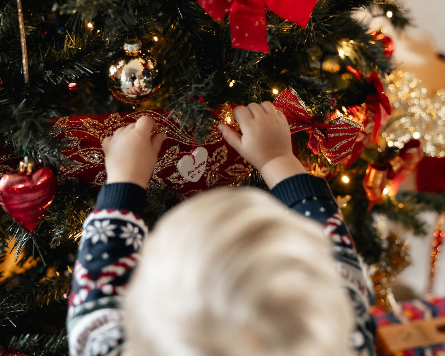 Young child in festive sweater decorating a Christmas tree with red ribbons and ornaments.