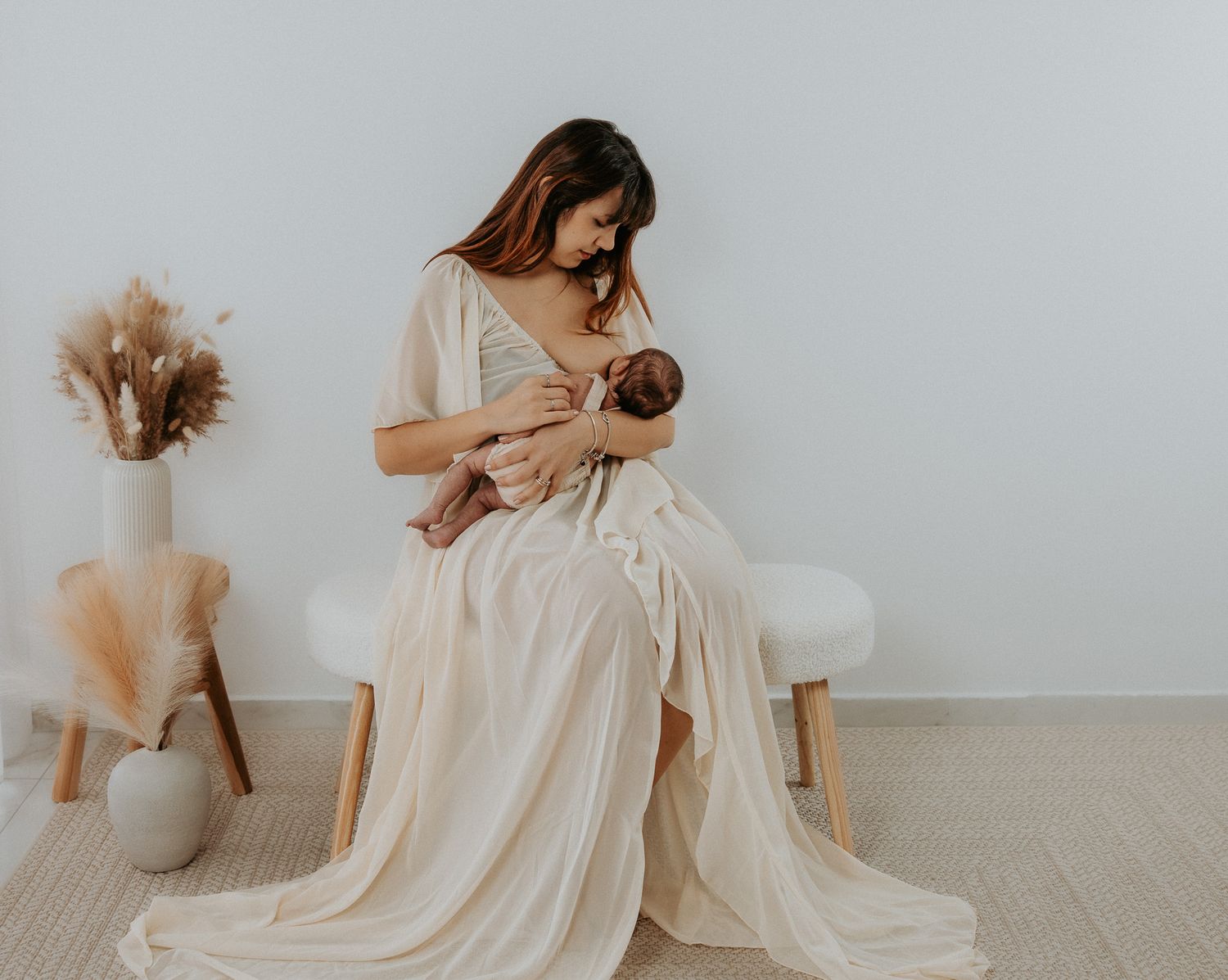 A person in a flowing cream dress sits on a chair nursing a baby, with dried pampas grass decor in the background.