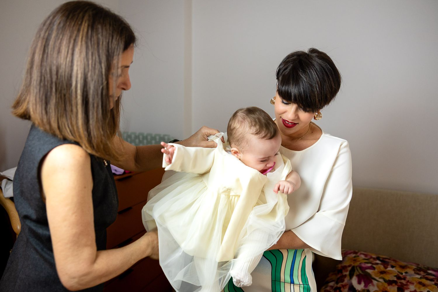 Two women admiring a baby dressed in a cream christening gown during what appears to be a baptism preparation.
