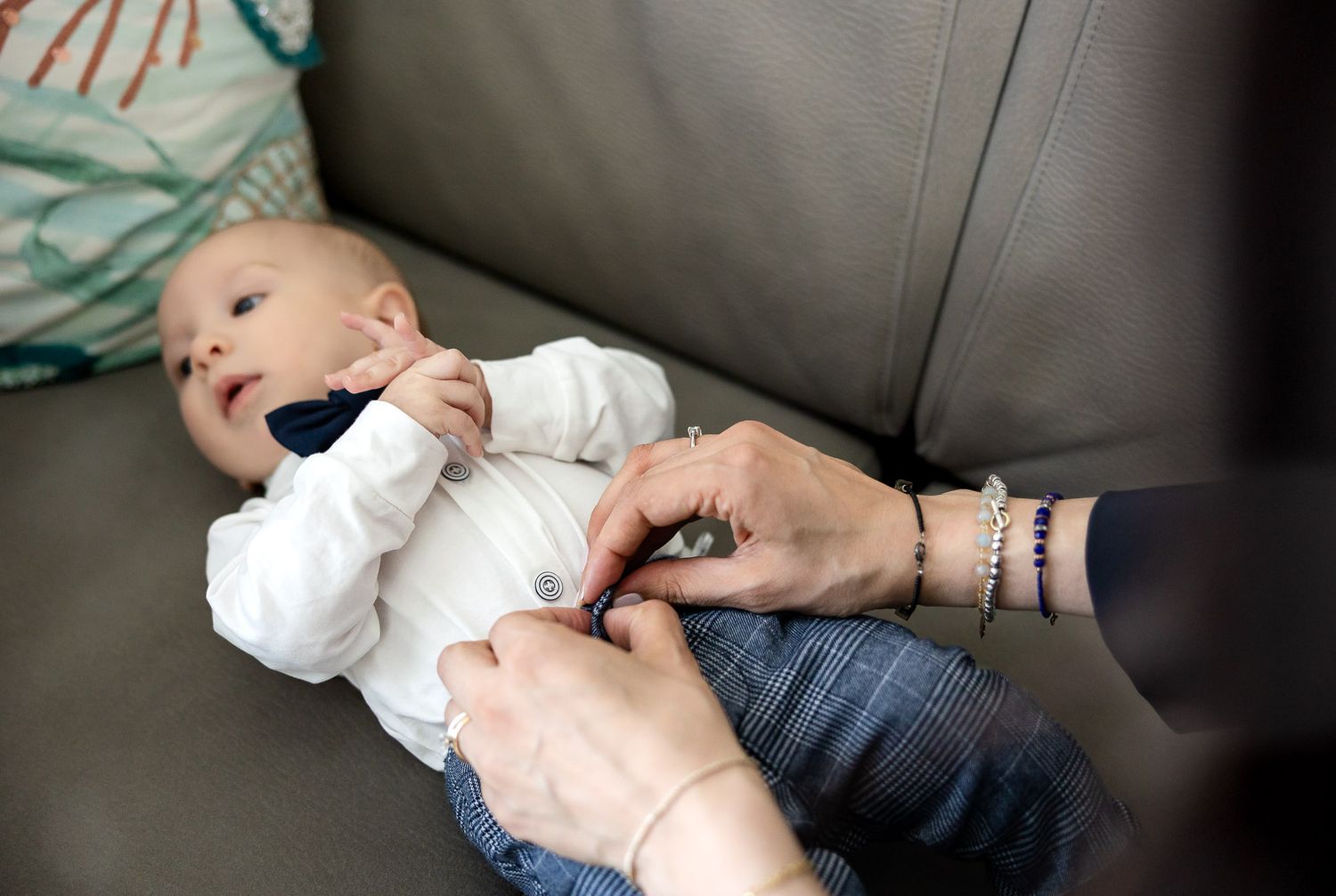 Adult hands fastening buttons on a white onesie worn by a newborn baby lying on a gray sofa.
