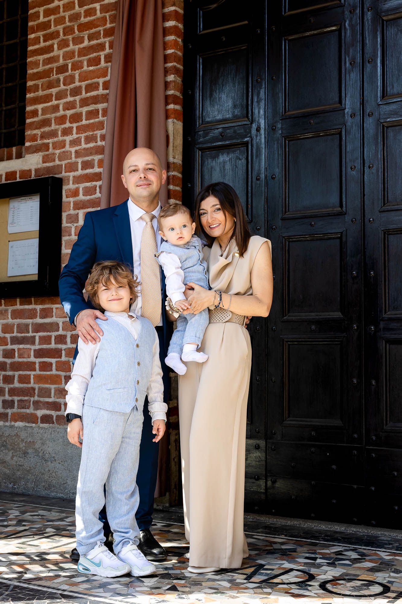 Family of four posing elegantly outside a church, parents dressed formally with two young children on a sunny day.