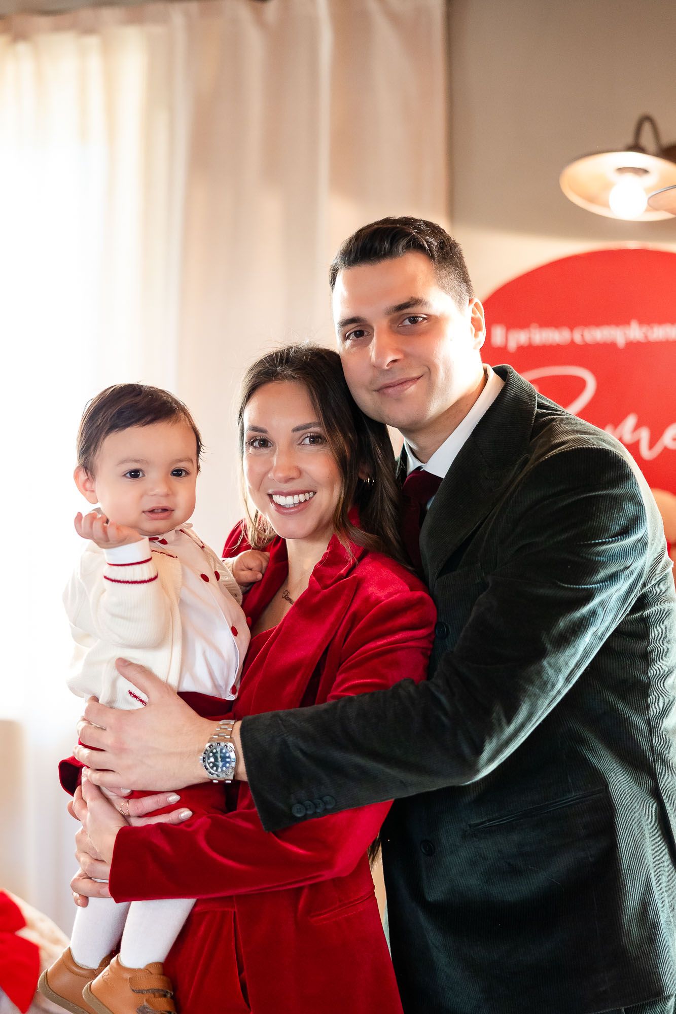 Smiling couple in red and green outfits holding their baby at a first birthday party celebration.