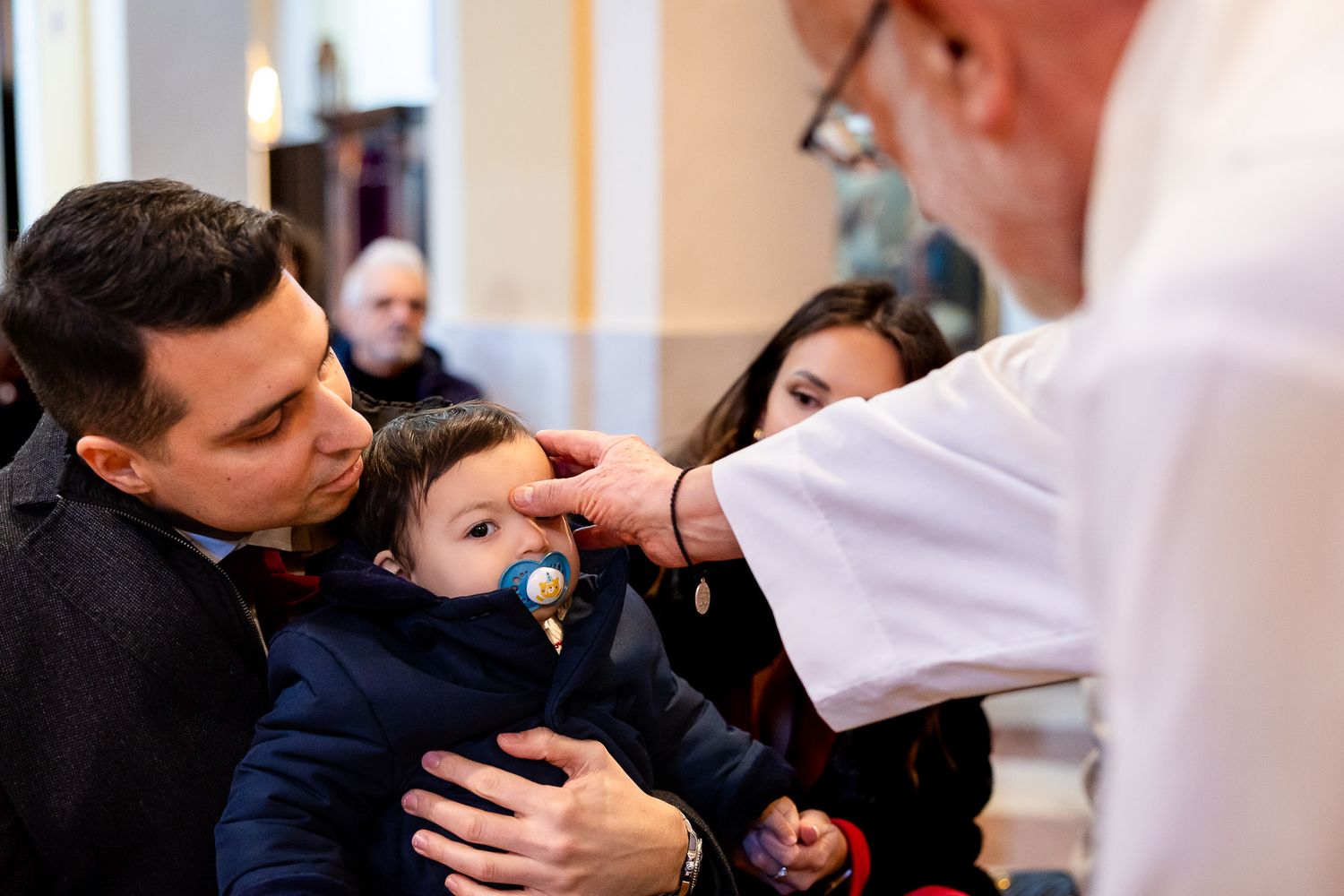 Priest anointing a baby with holy oil during a Catholic baptism ceremony while father holds the infant.