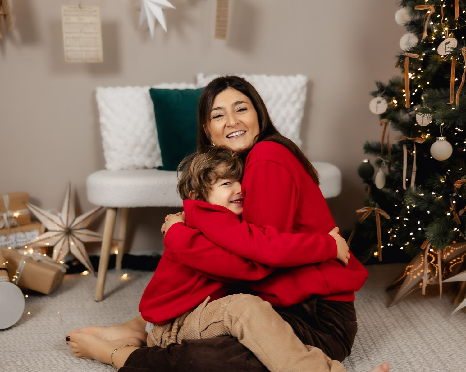 A woman in a red sweater joyfully hugs a young boy near a decorated Christmas tree with gifts.