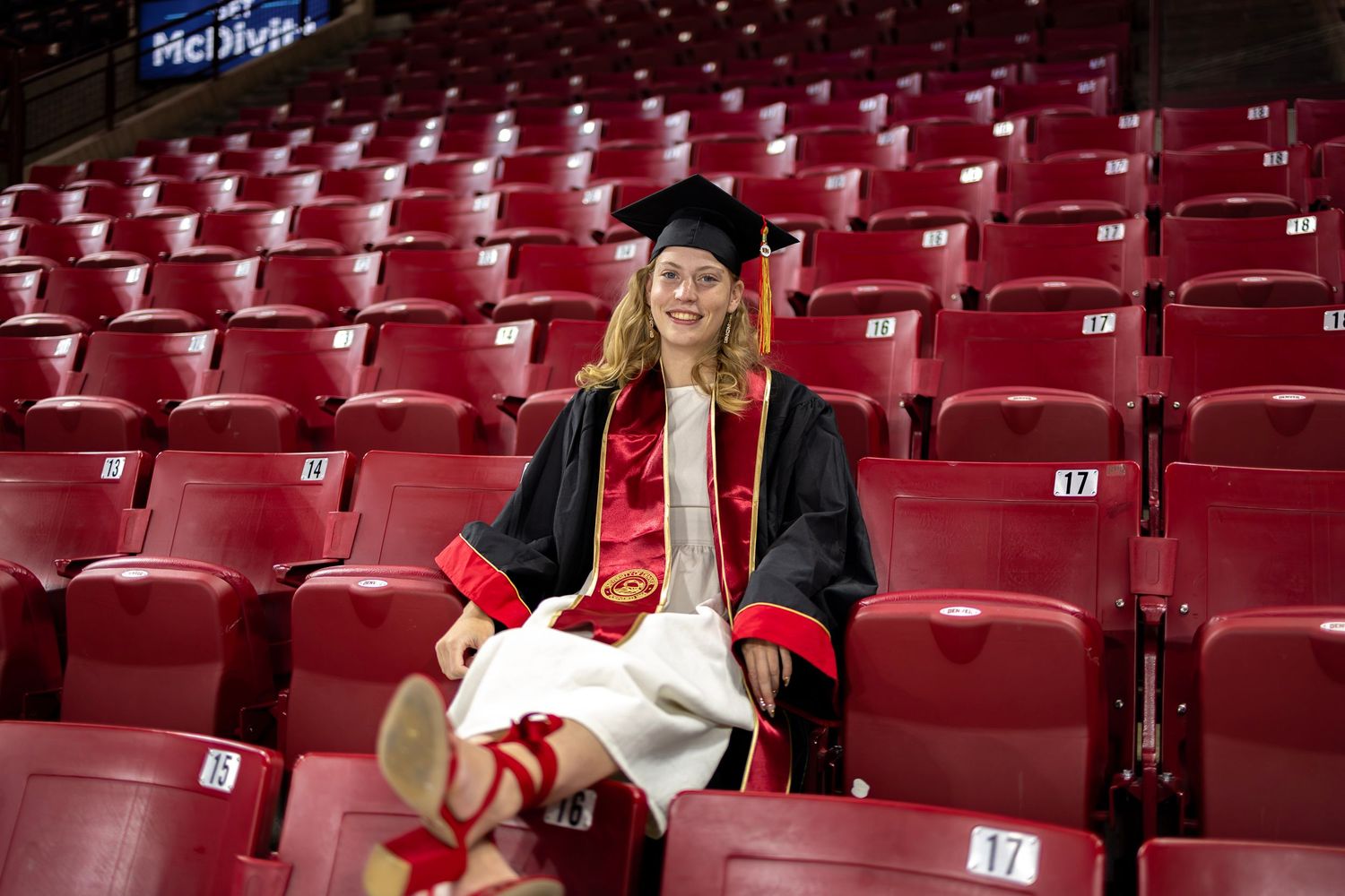 A smiling graduate in cap and gown sits among red stadium seats, wearing a red sash and holding a diploma.