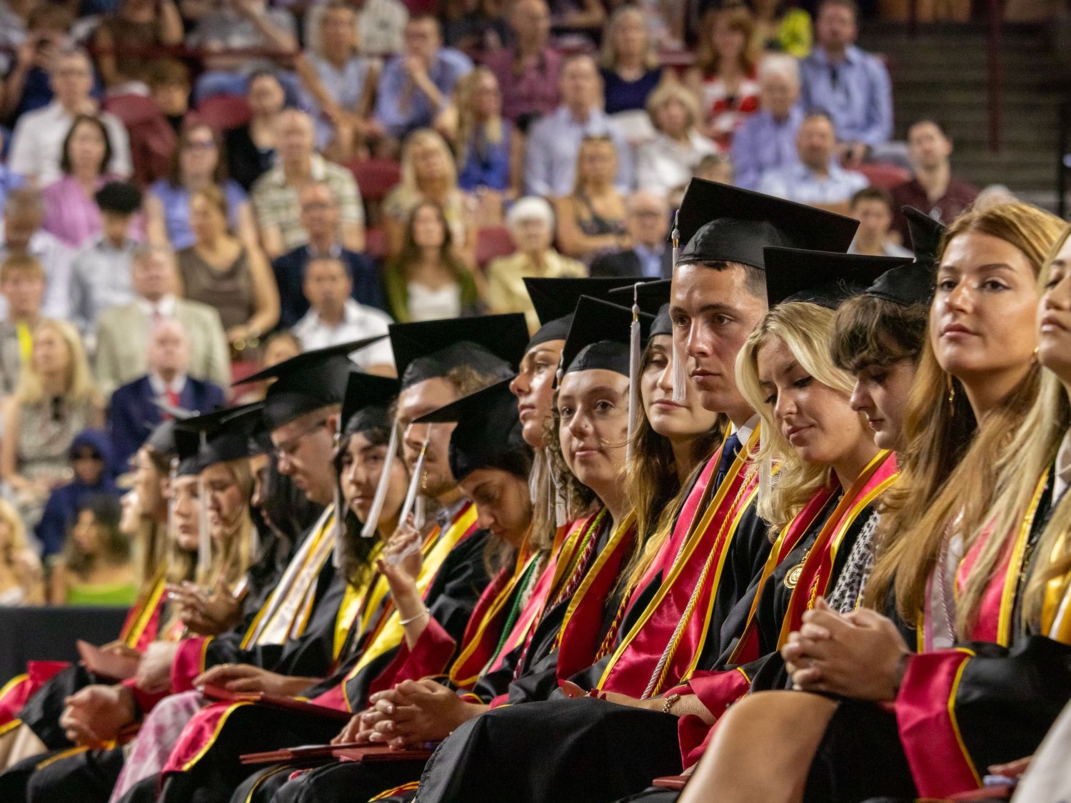 Graduates wearing black caps and colorful stoles sit together during commencement ceremony in an auditorium.