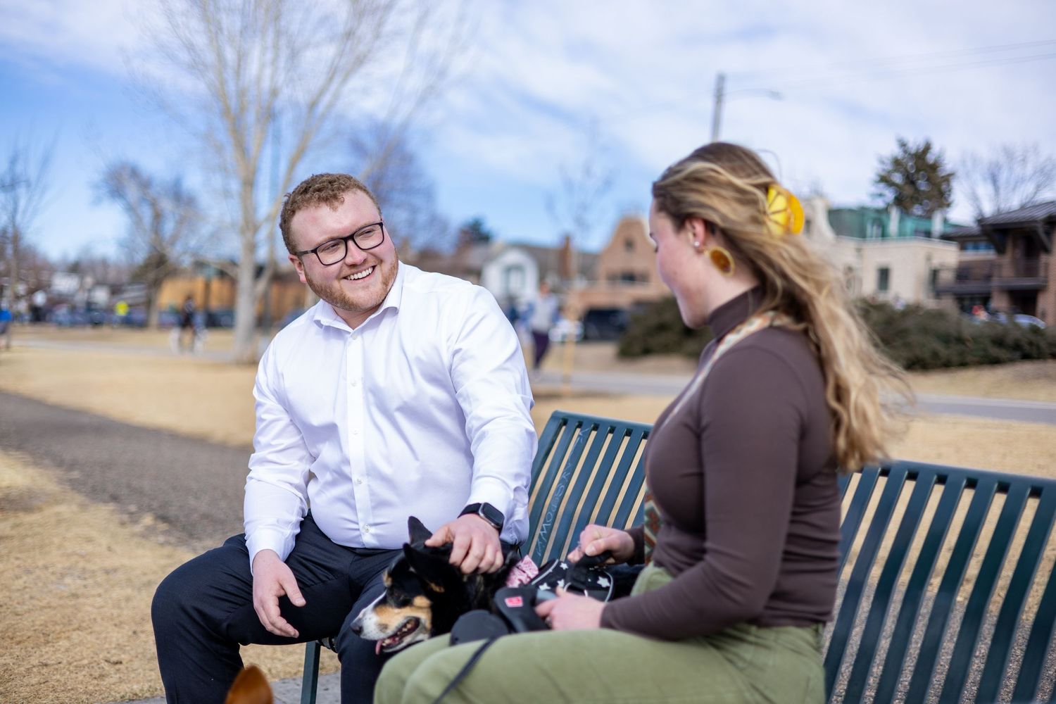A couple sits together on a park bench during an outdoor photo session on a sunny day.