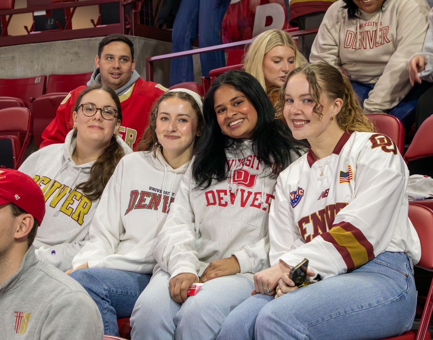 Four smiling University of Denver students pose together in DU sweatshirts and jerseys at a hockey game.