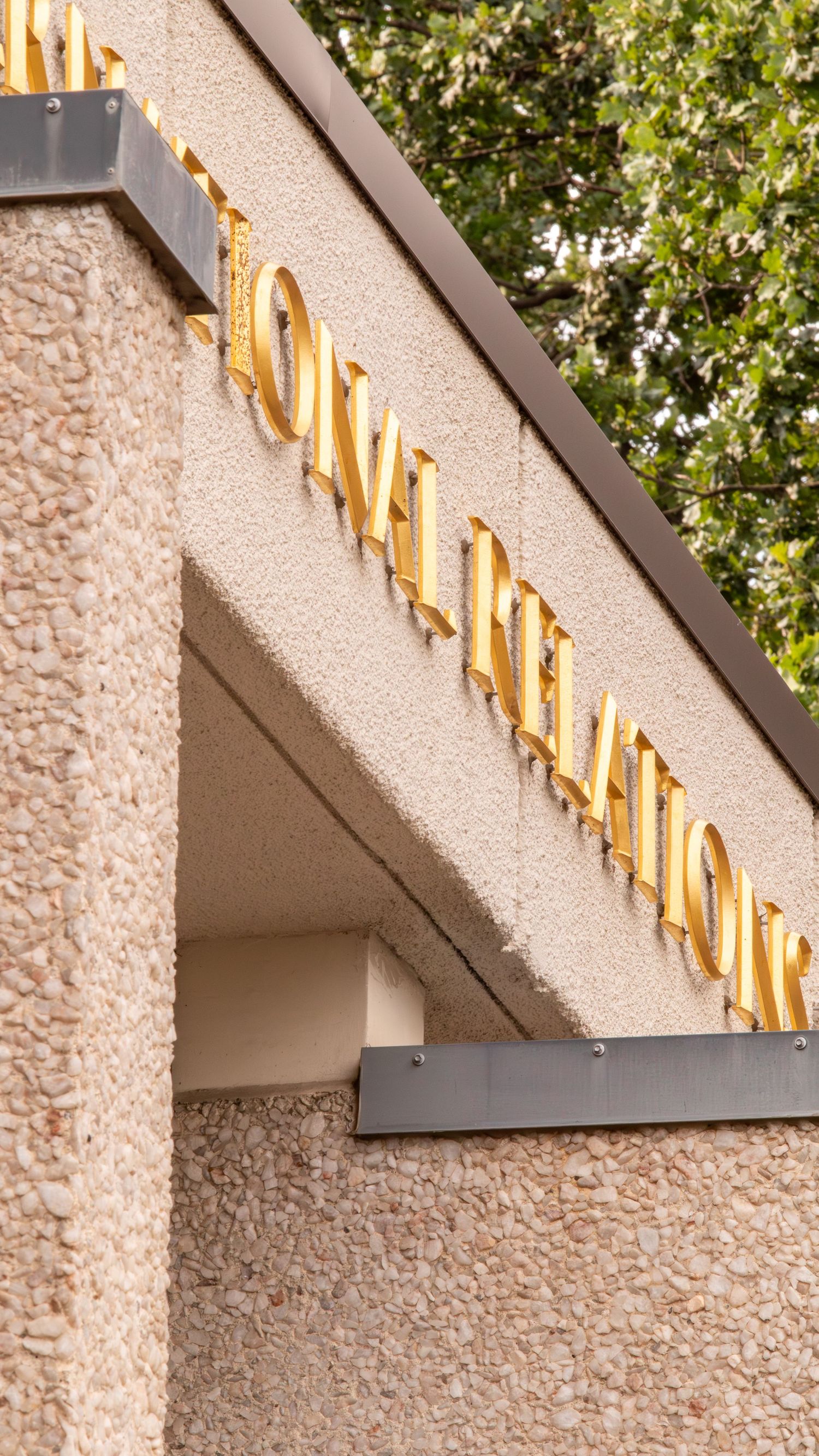 Gold letter signage reading Community Relations on a textured stone building exterior with trees in background.