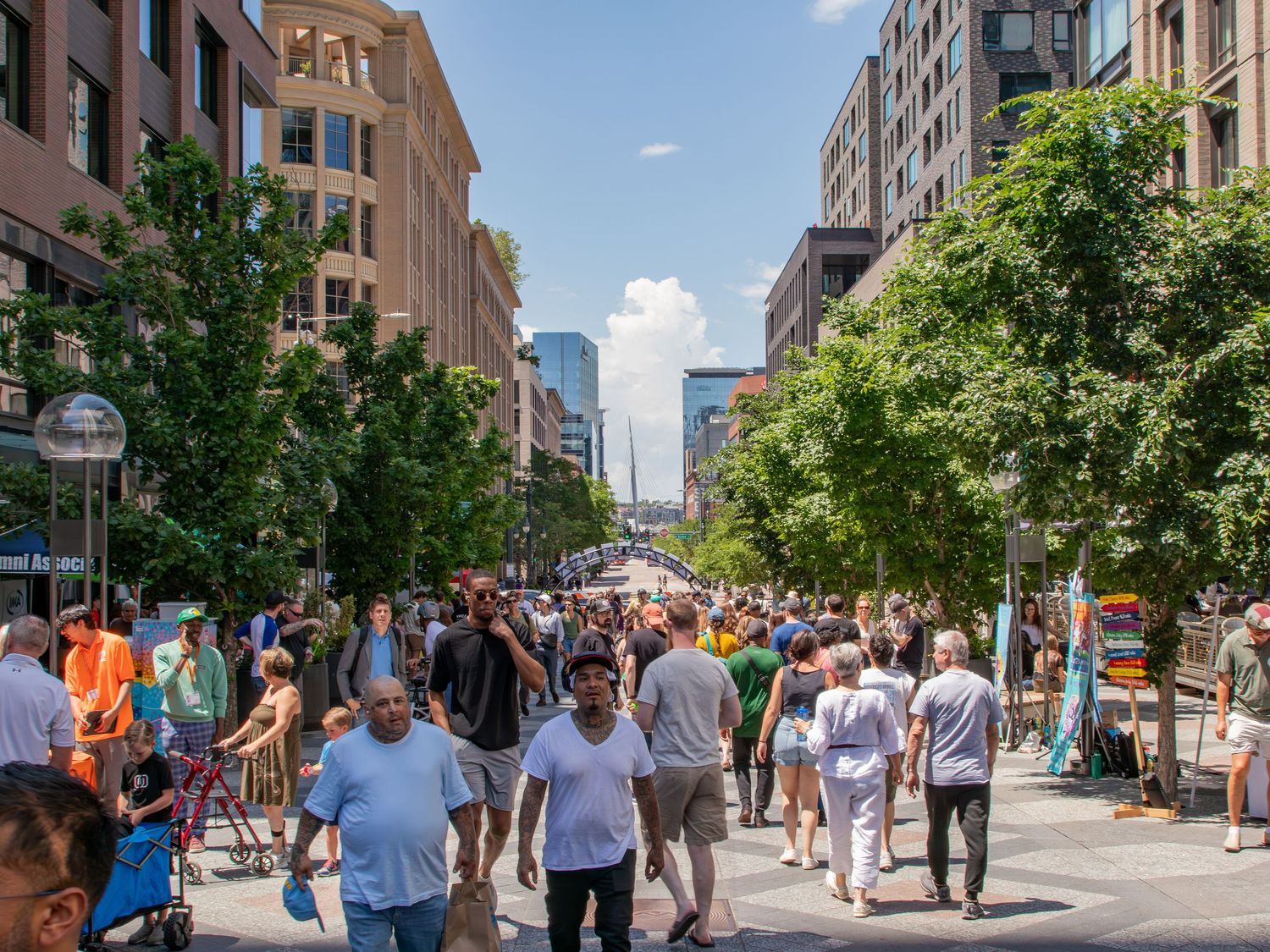 Crowds of people walk along a busy urban pedestrian street lined with trees and modern buildings on a sunny day.