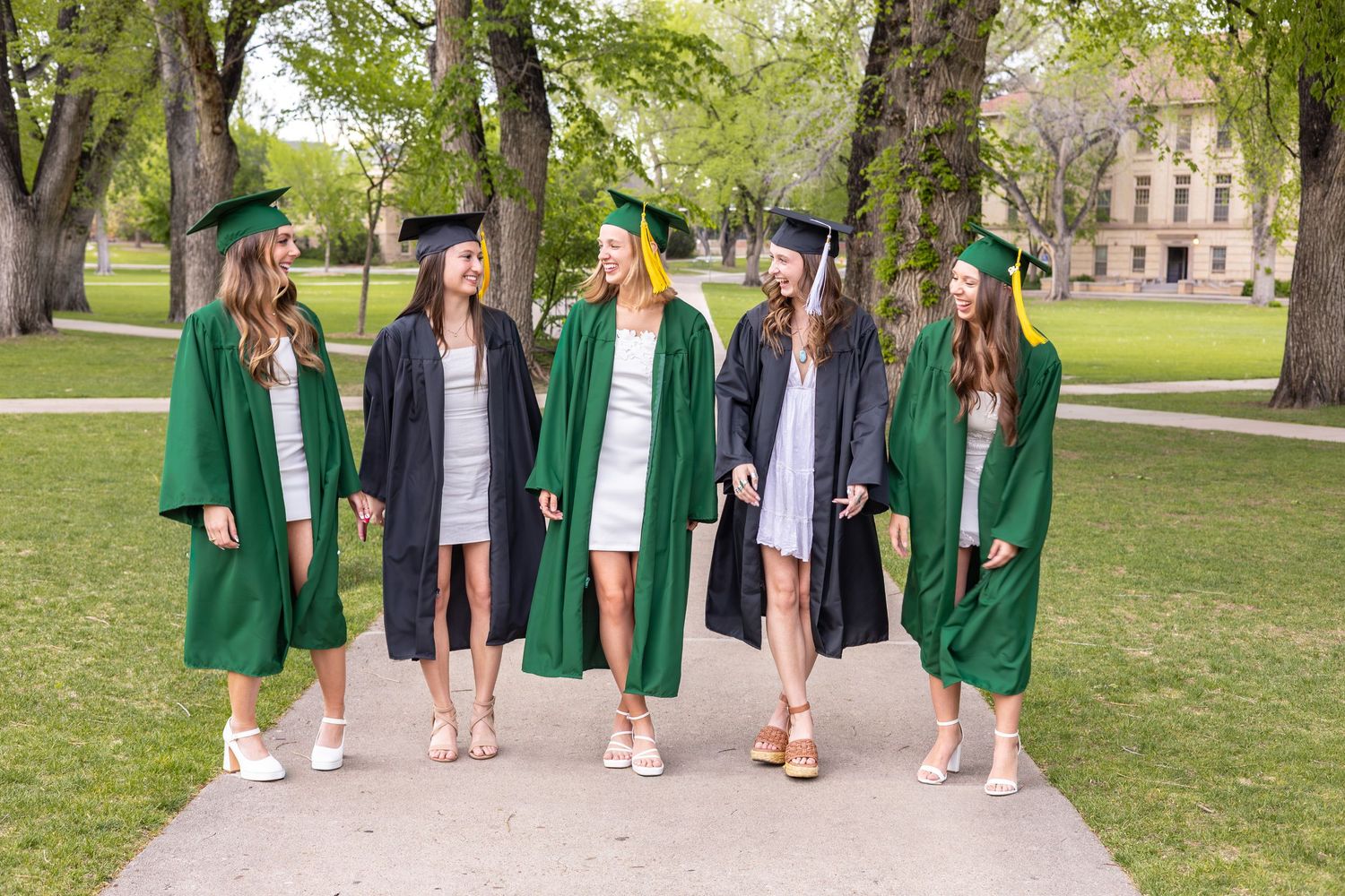 Five female graduates in green and black caps and gowns walk together on a campus pathway surrounded by trees.