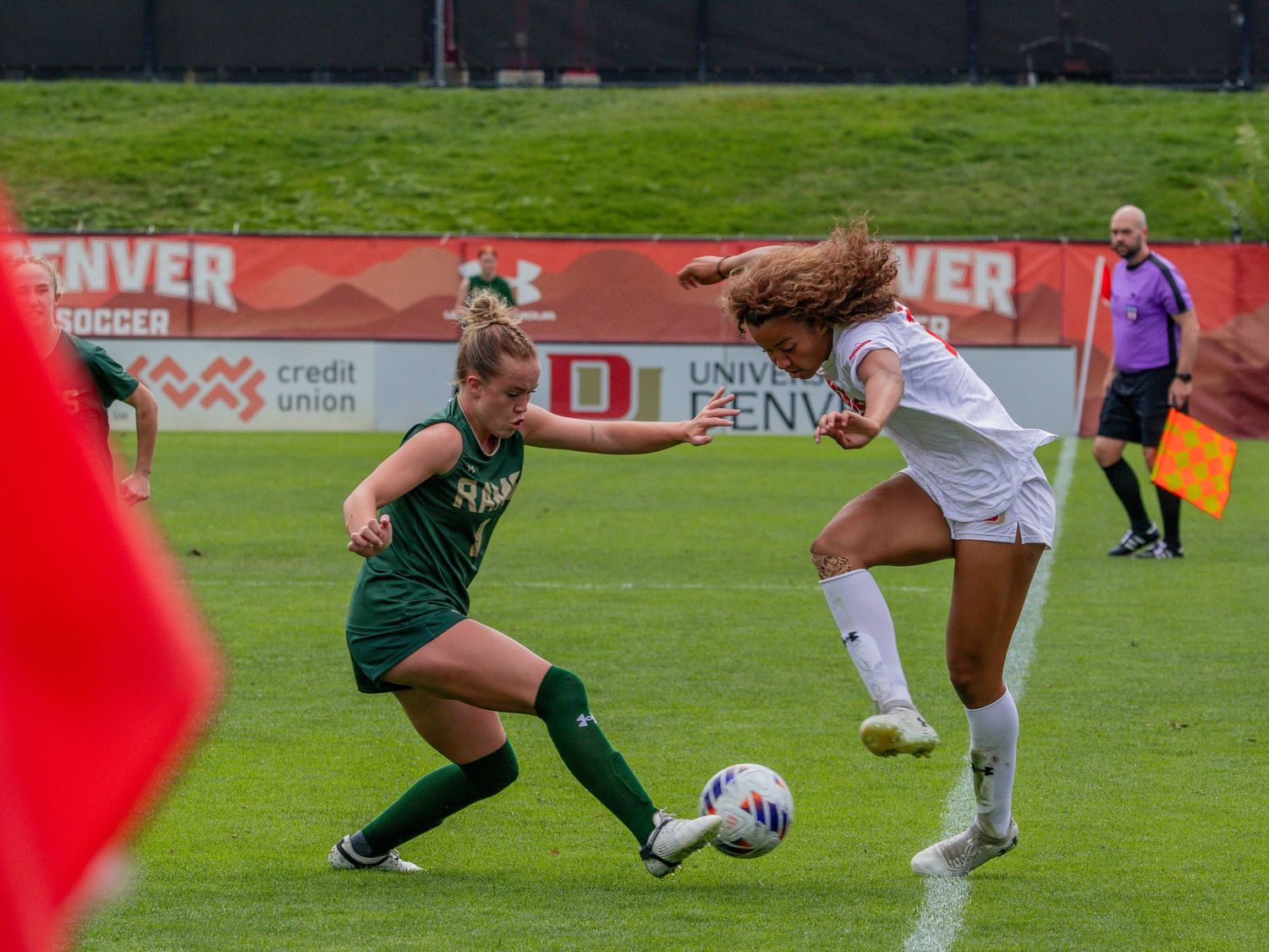 Two female soccer players compete for the ball during a University of Denver women's soccer match on a red turf field.