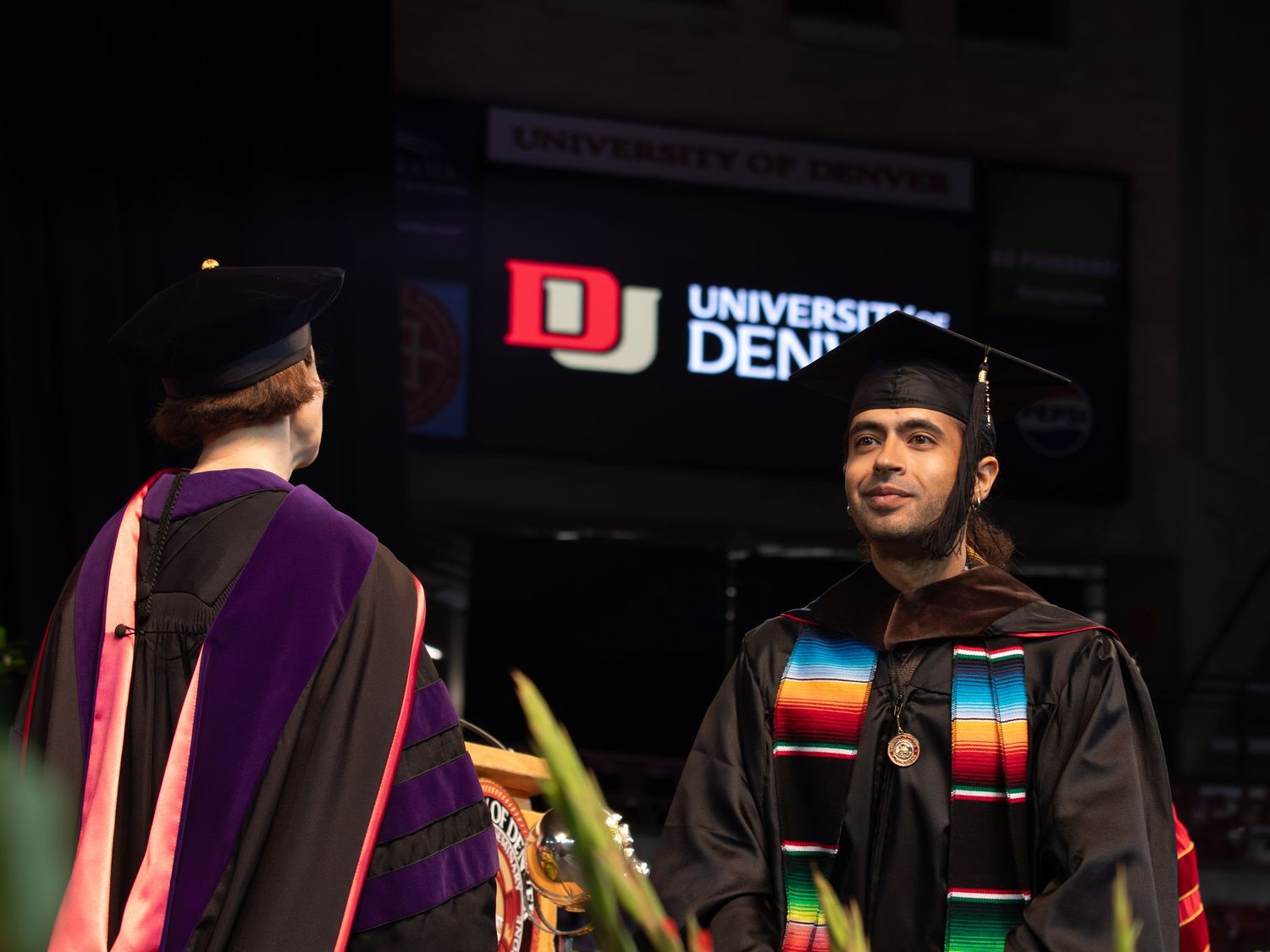 University of Denver graduation ceremony with two graduates in academic regalia and colorful stoles on stage.