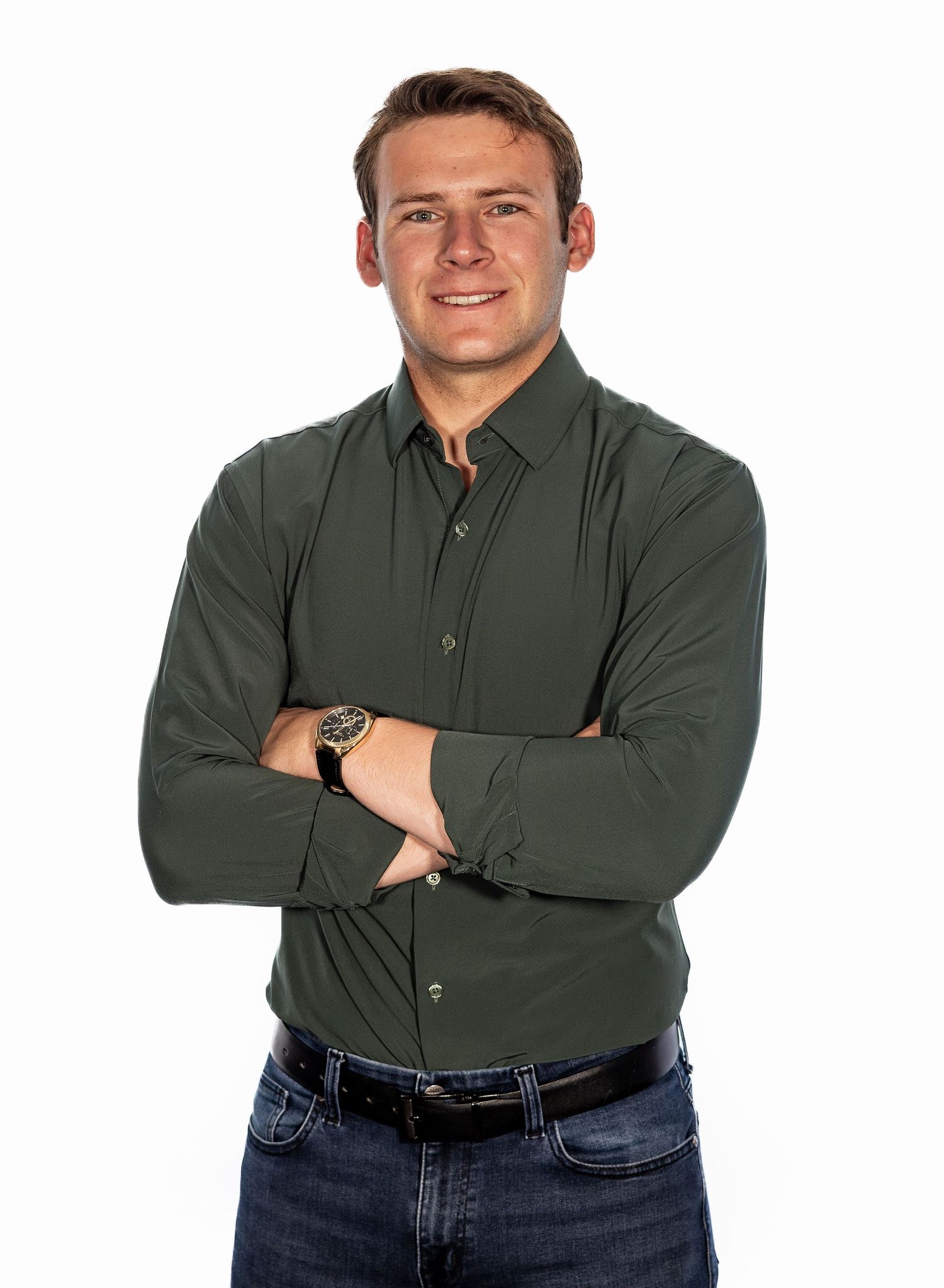 Young professional man in dark green button-up shirt with arms crossed, smiling confidently against white background.