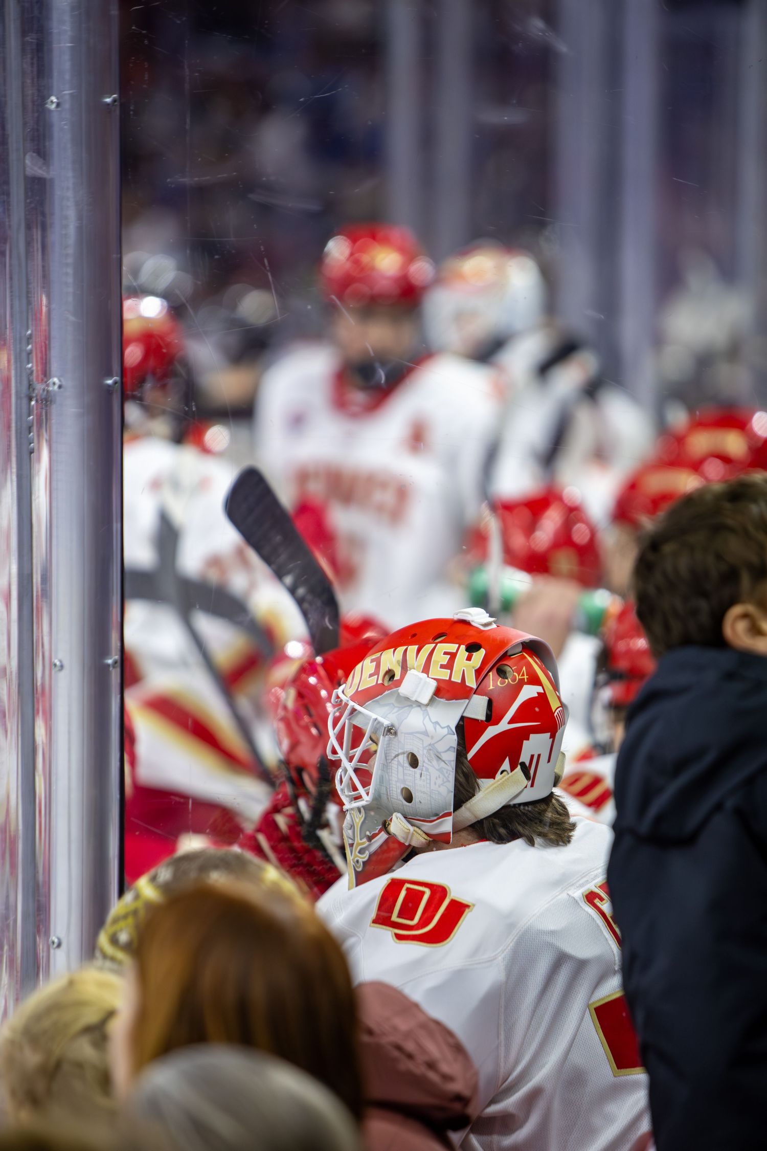 Denver hockey goalie in red helmet sits on bench during game.