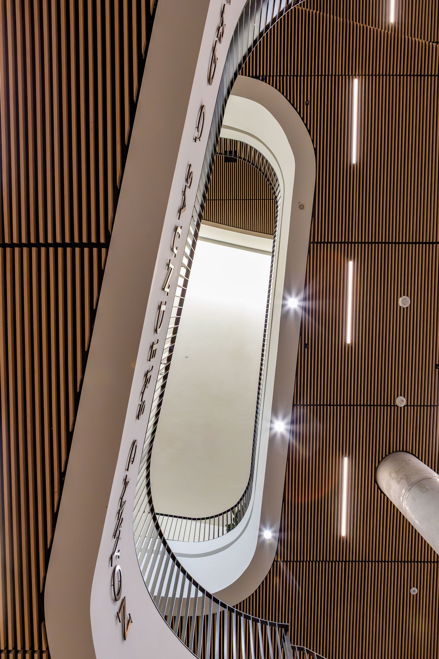 Modern spiral staircase viewed from below, featuring sleek white railings and warm wood-paneled walls with elegant lighting.