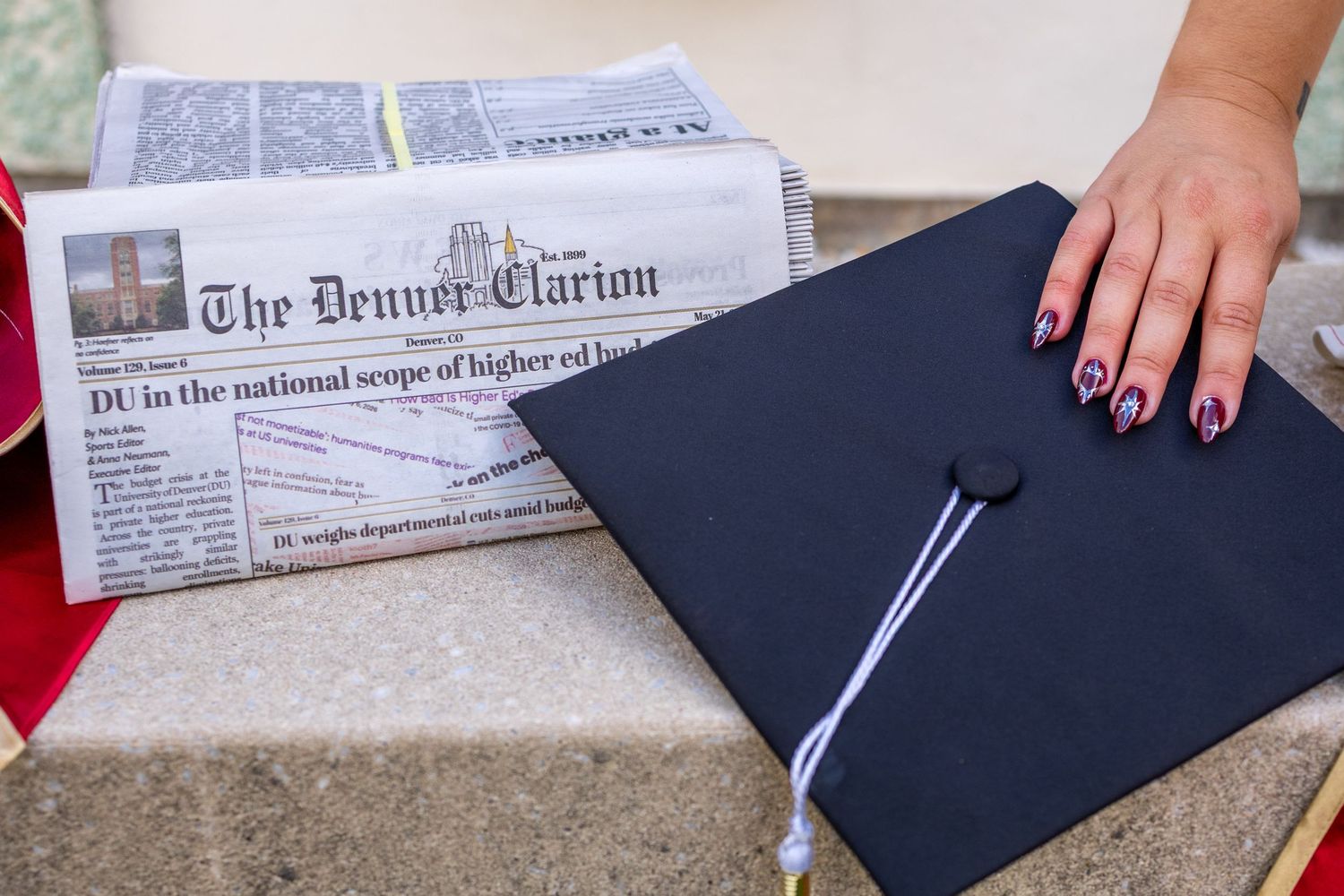 Graduation cap and The Denver Clarion newspaper on stone steps, with a hand displaying patriotic nail art.