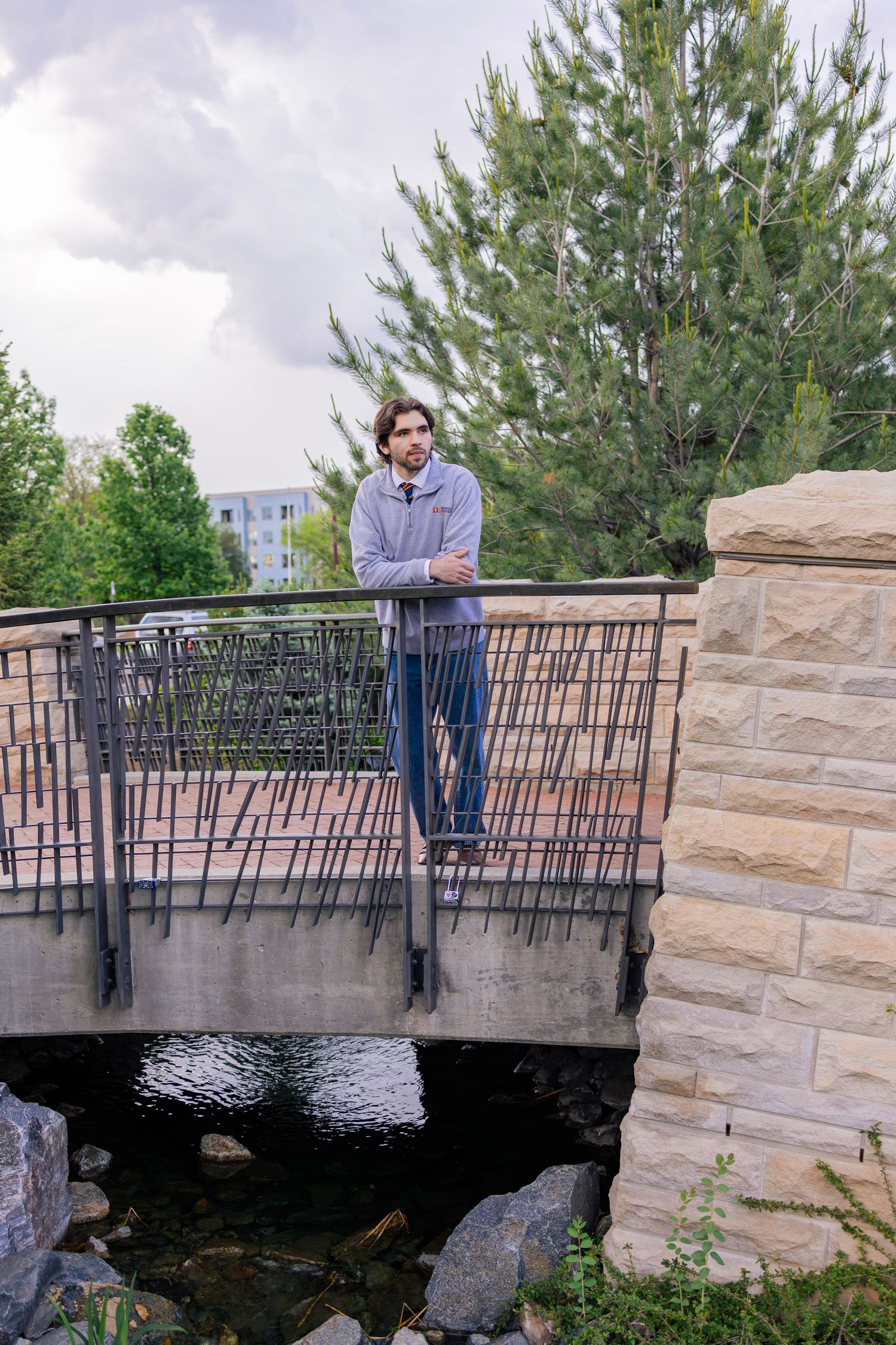 Man with long hair stands on a stone bridge with metal railings over a rocky stream, surrounded by trees.