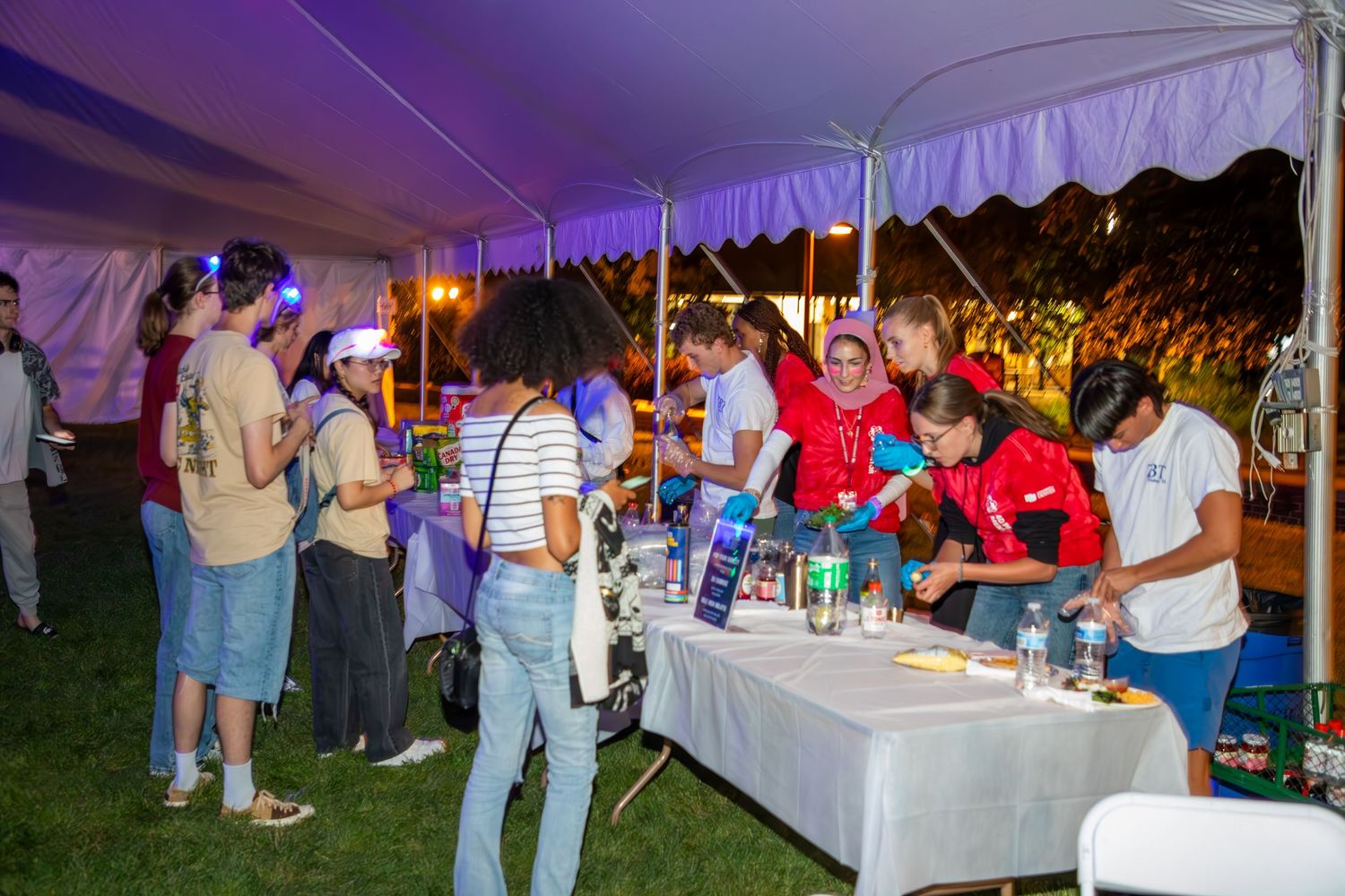 People gather under a large tent at an outdoor evening event, browsing tables with colorful displays and drinks.