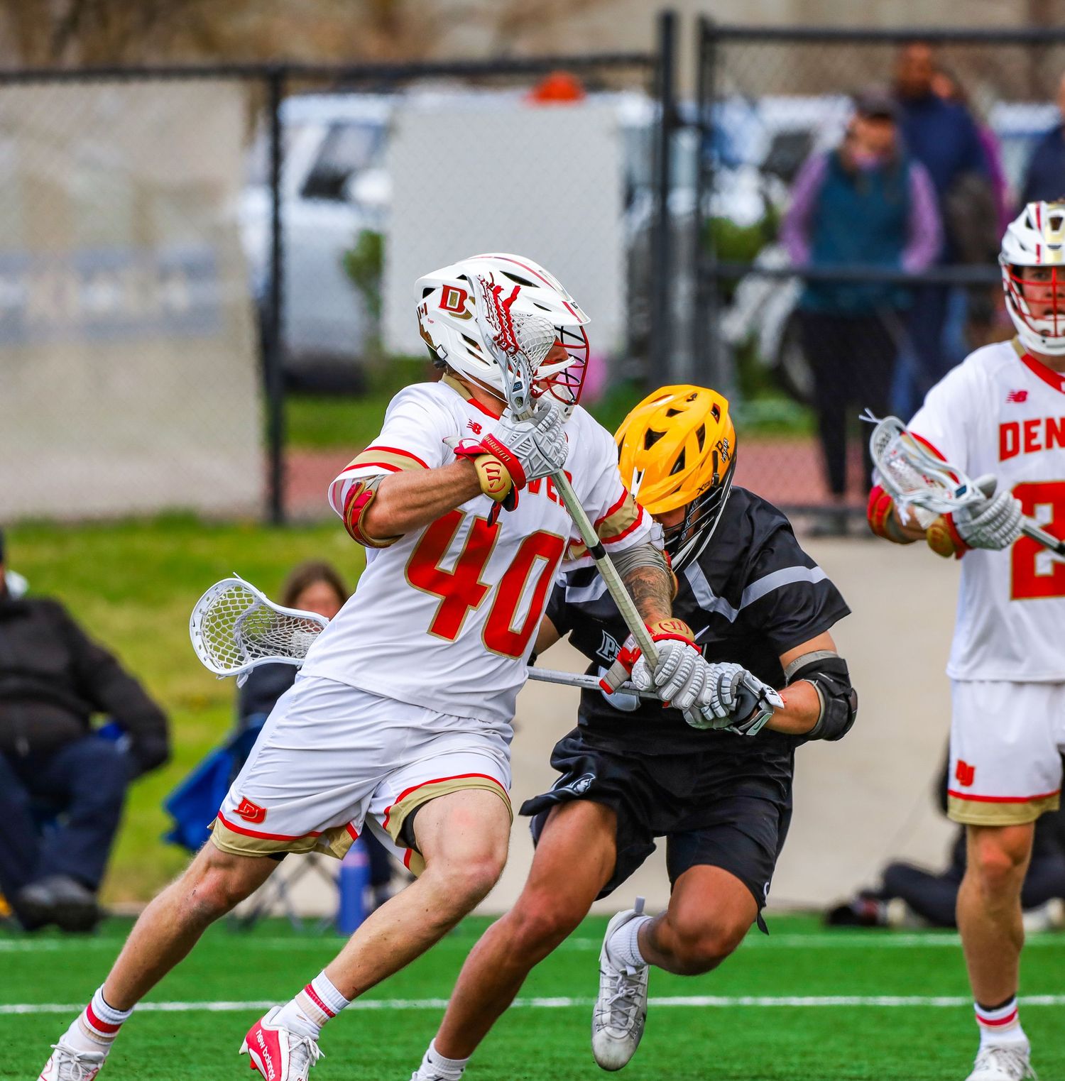 Lacrosse players in red and white uniforms compete against opponents in black during an intense match on a grass field.
