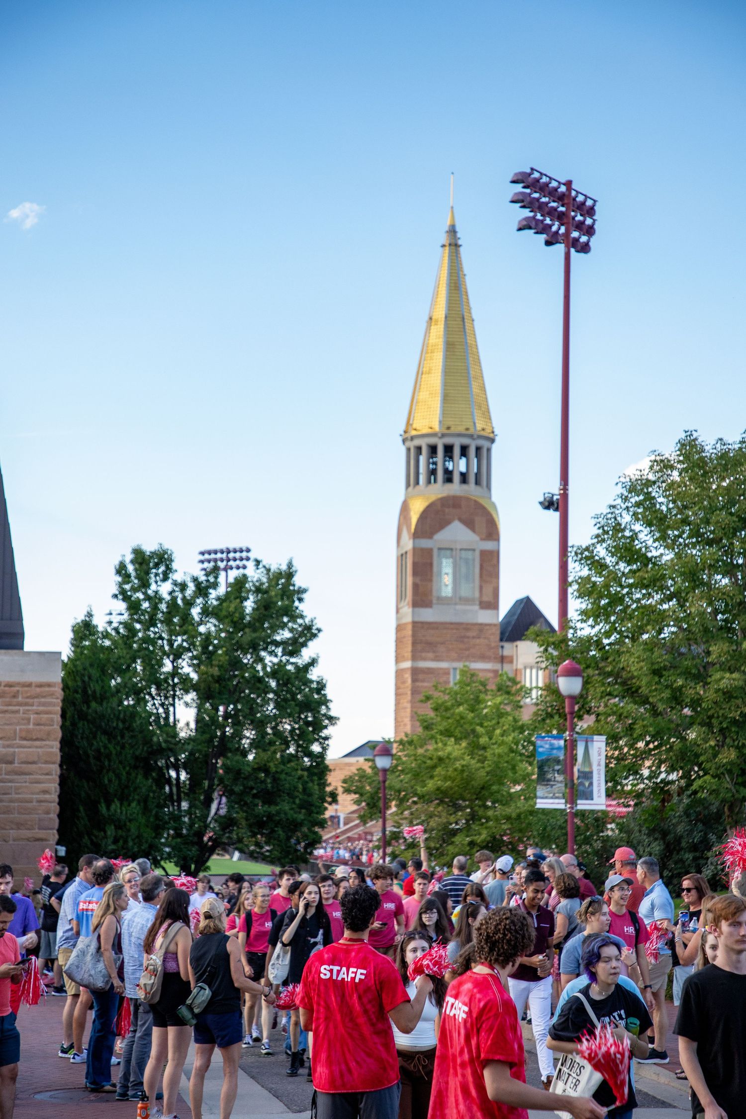 Crowd of students and staff gather on a university campus plaza with a historic gold-spired bell tower in the background.