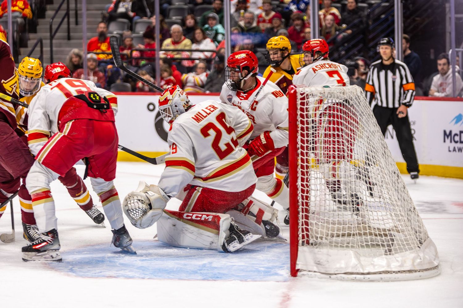 College hockey goalie in red jersey defends net during intense game action on ice rink.