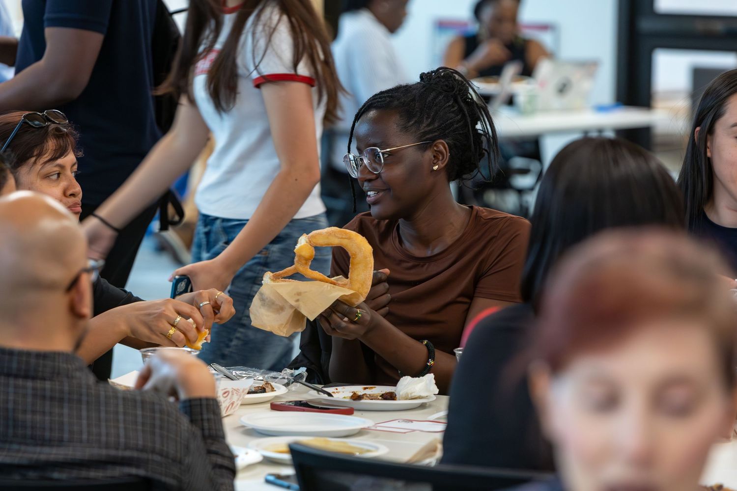 A smiling woman holds a pretzel at a busy indoor gathering with diverse groups of people socializing around tables.