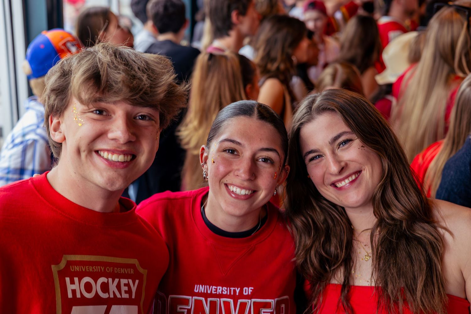 Three smiling students in red University of Denver hockey shirts pose together at a campus sporting event.