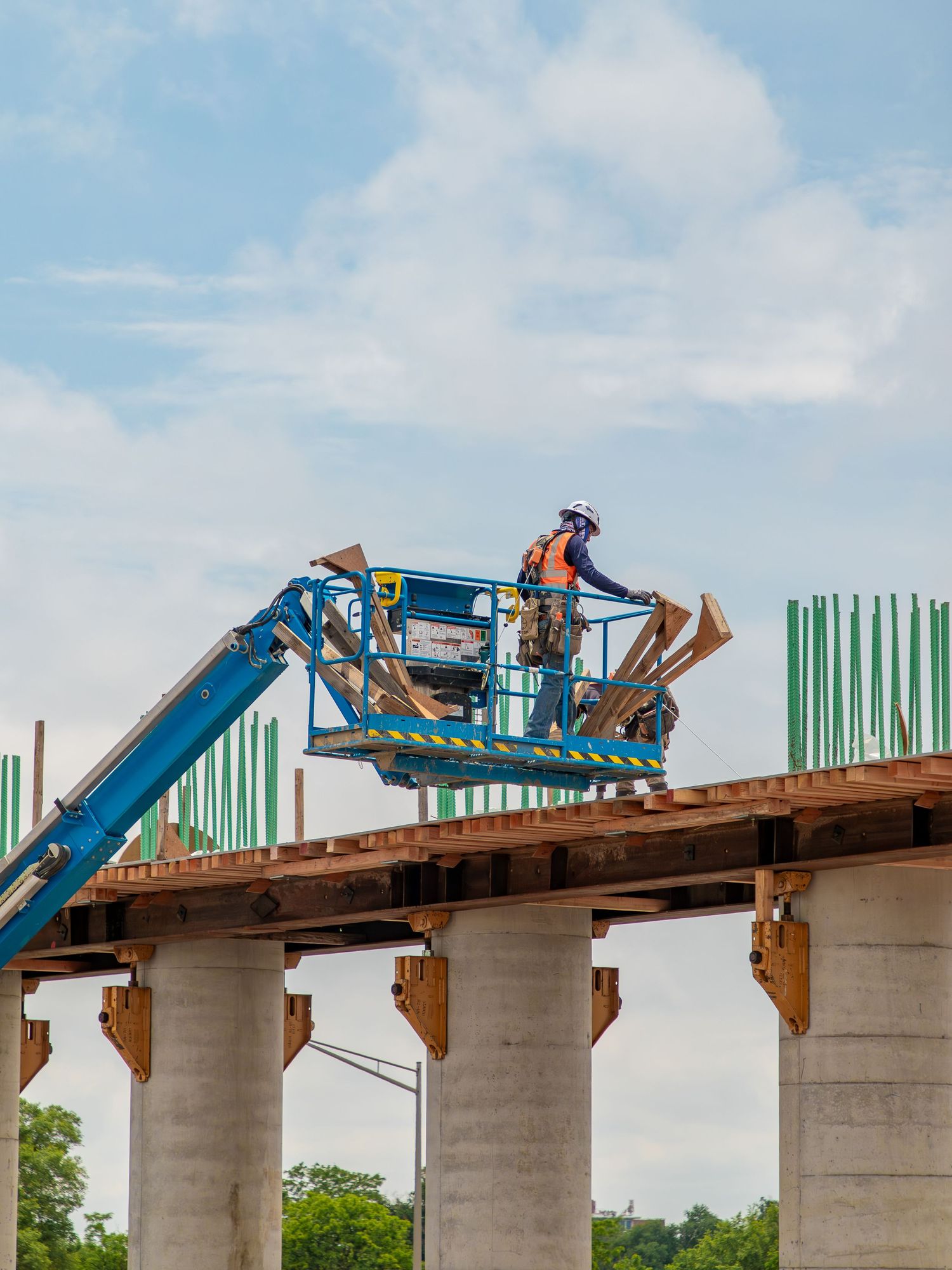 Construction crews work on elevated concrete bridge pillars with blue machinery and equipment.