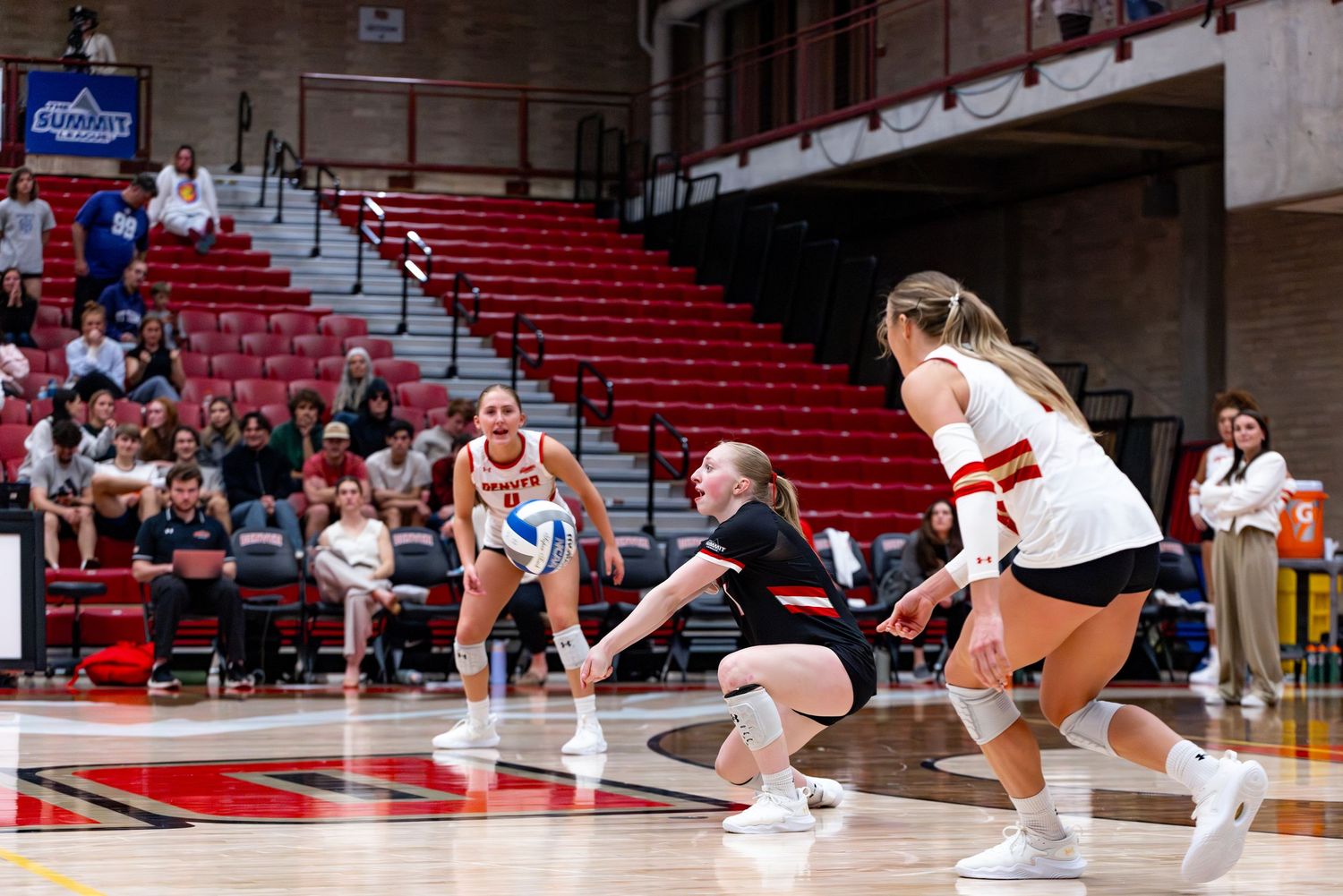 Female volleyball players in red and white uniforms compete indoors, one player diving to dig the ball during an intense match.