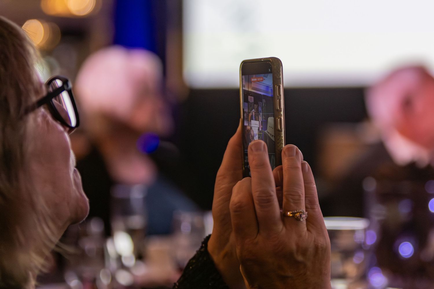 A person holds up a smartphone to record video at a dimly lit indoor event with blurred lights in the background.