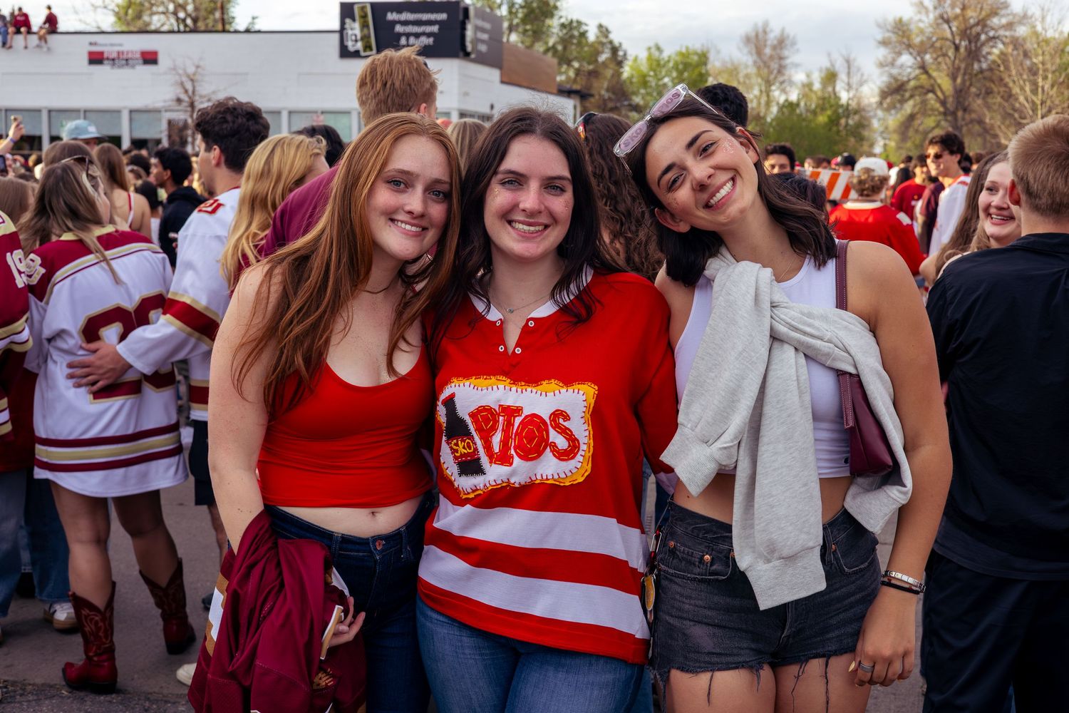 Three young women in red outfits smiling together at an outdoor sports fan event.