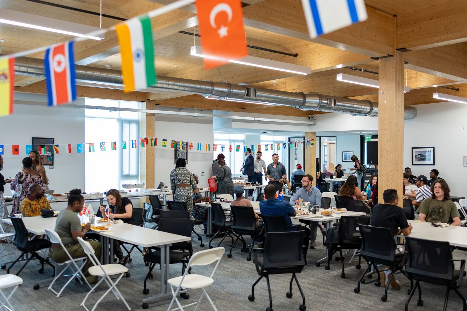 Busy open workspace with international flags hanging from ceiling, people working at tables in modern office.