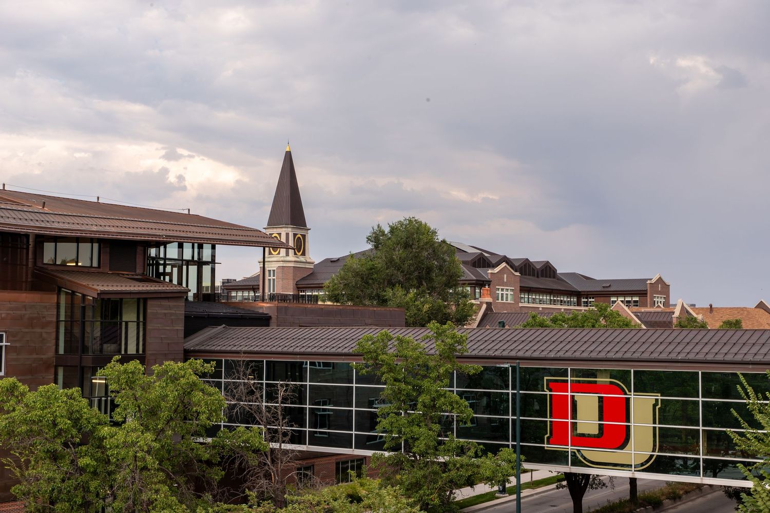 University of Denver campus buildings with iconic red D logo and church steeple against cloudy sky.
