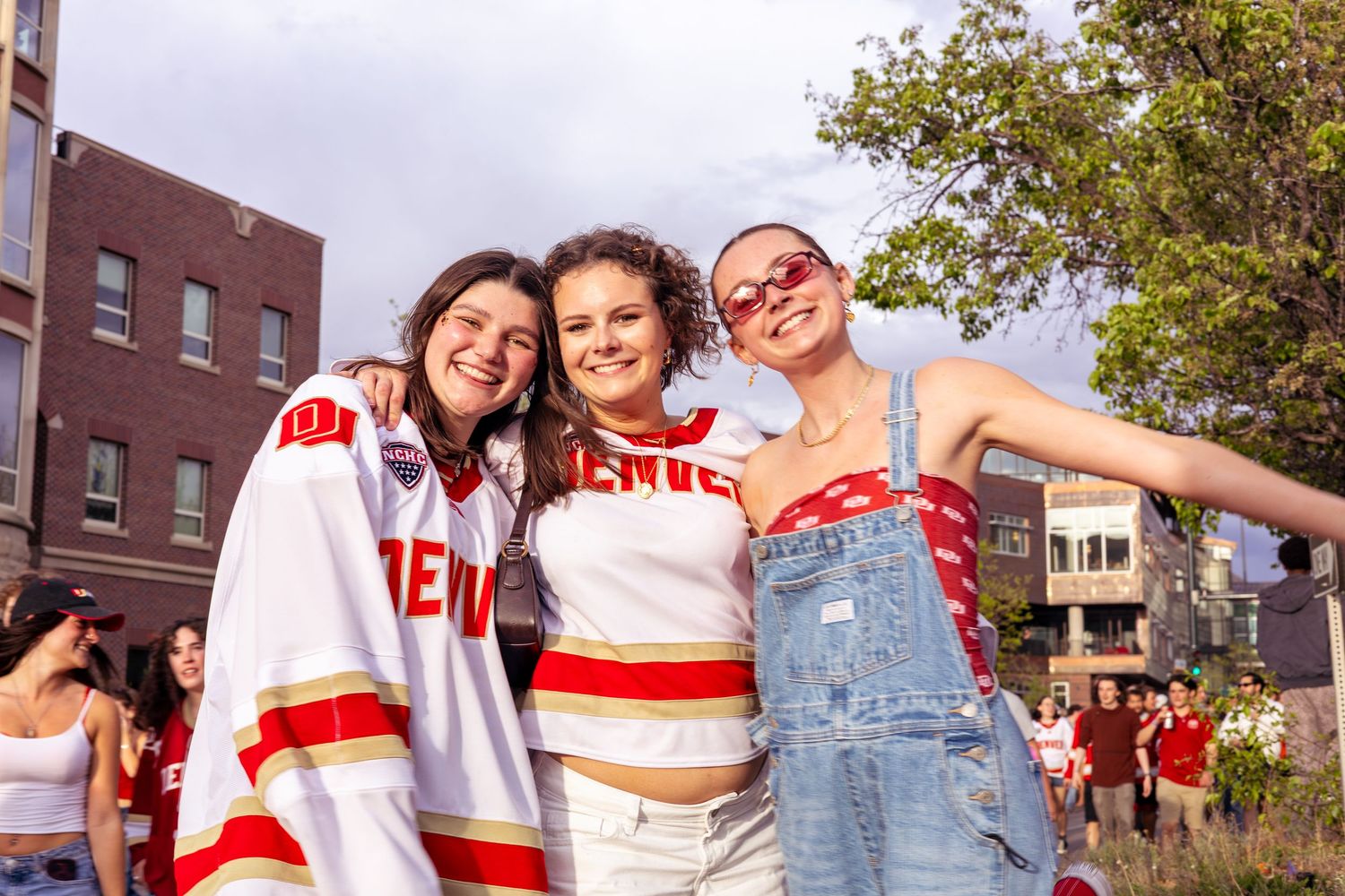 Three smiling women in red and white Denver sports gear pose together outdoors at a fan event.