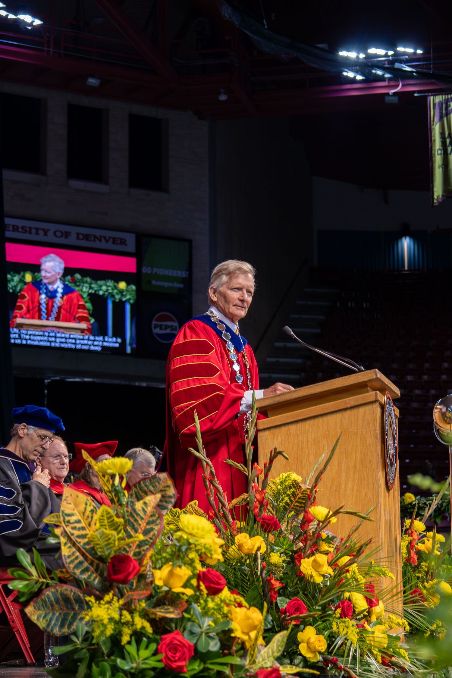University of Denver chancellor speaks at graduation ceremony podium in red academic regalia surrounded by colorful flowers.