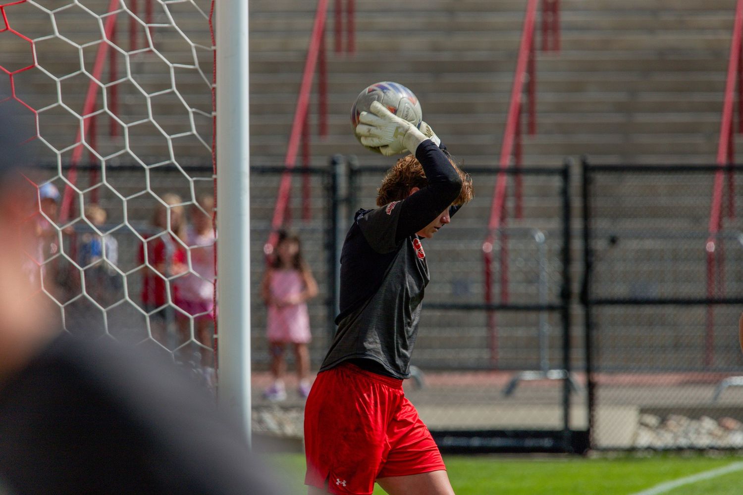 Soccer goalkeeper in red shorts and black jersey catching a ball in front of the net during a match.