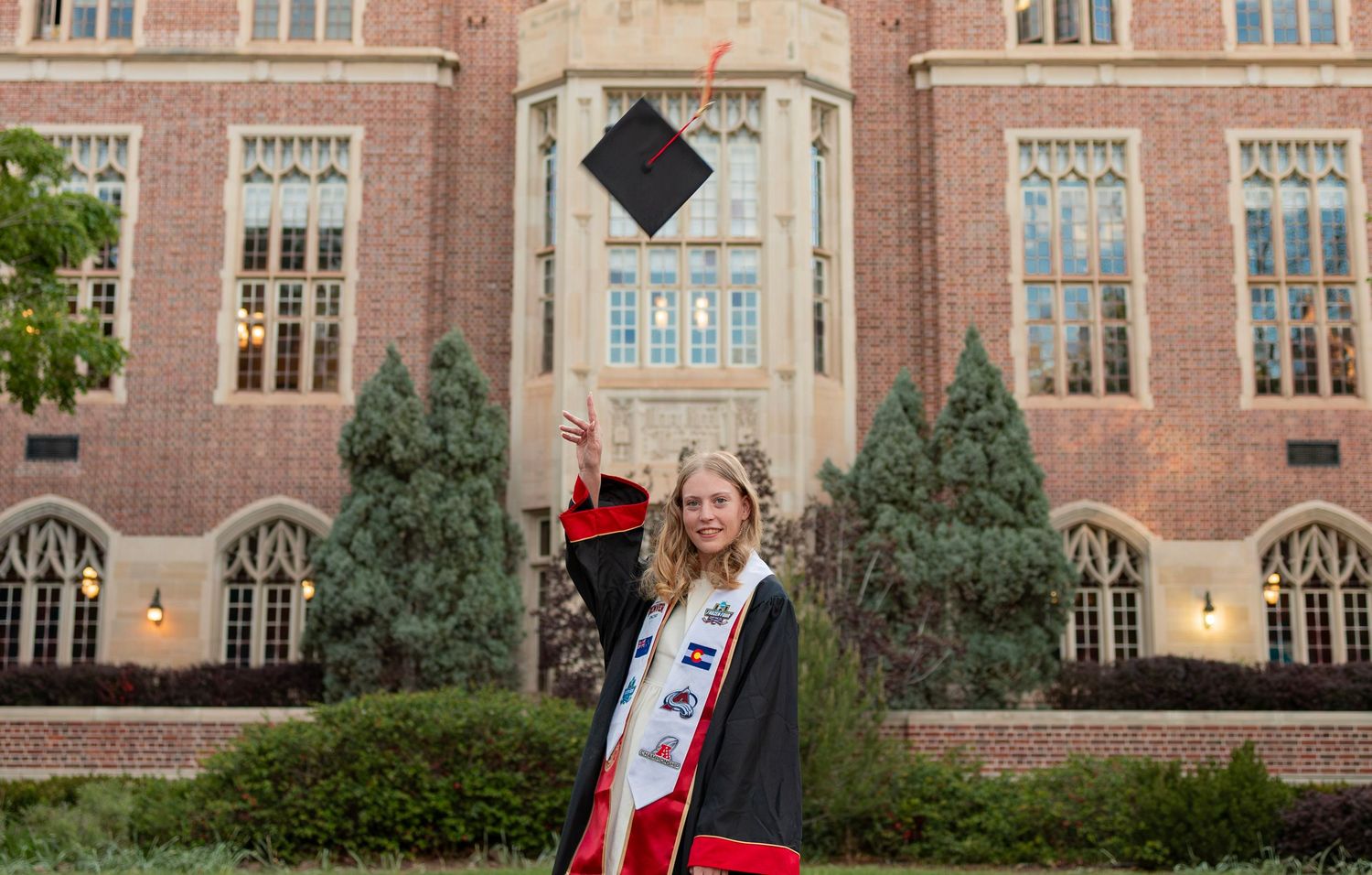 Graduate tosses cap in front of brick university building wearing cap, gown, and graduation stole.