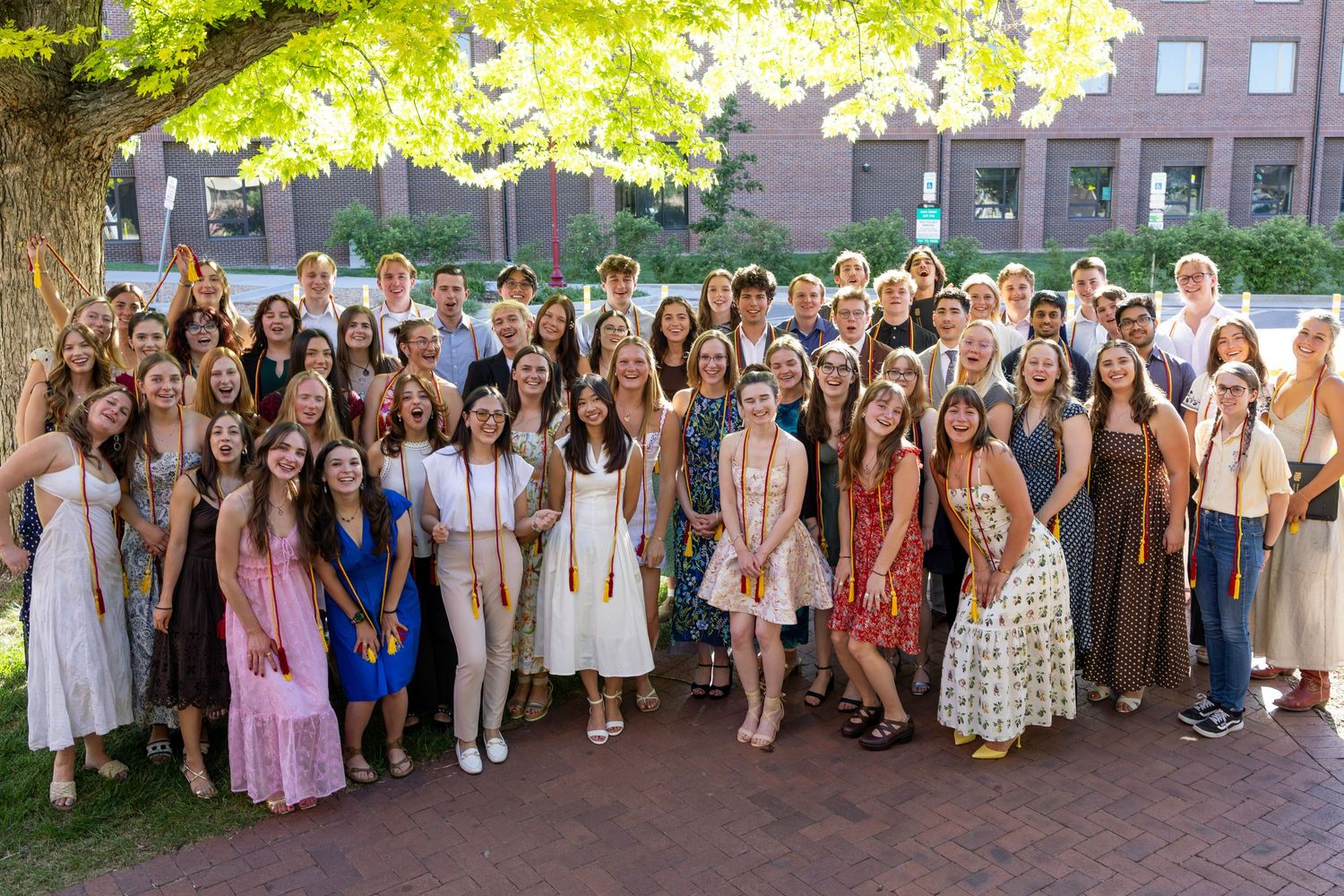 Large group of college graduates in summer attire posing outdoors near brick building with lush green trees.