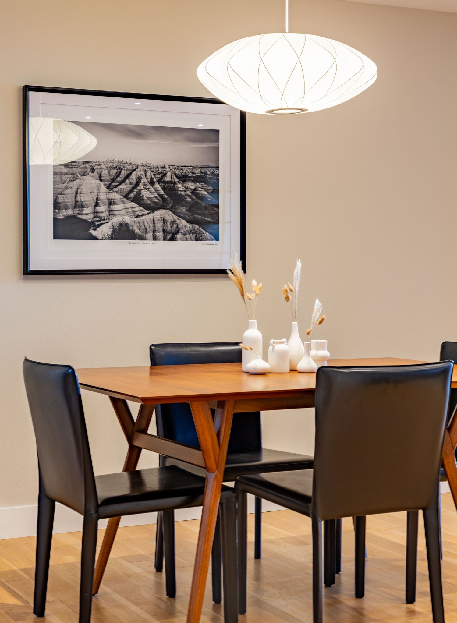 Modern dining area with wooden tables, black leather chairs, white pendant lamps and framed landscape artwork on beige walls.