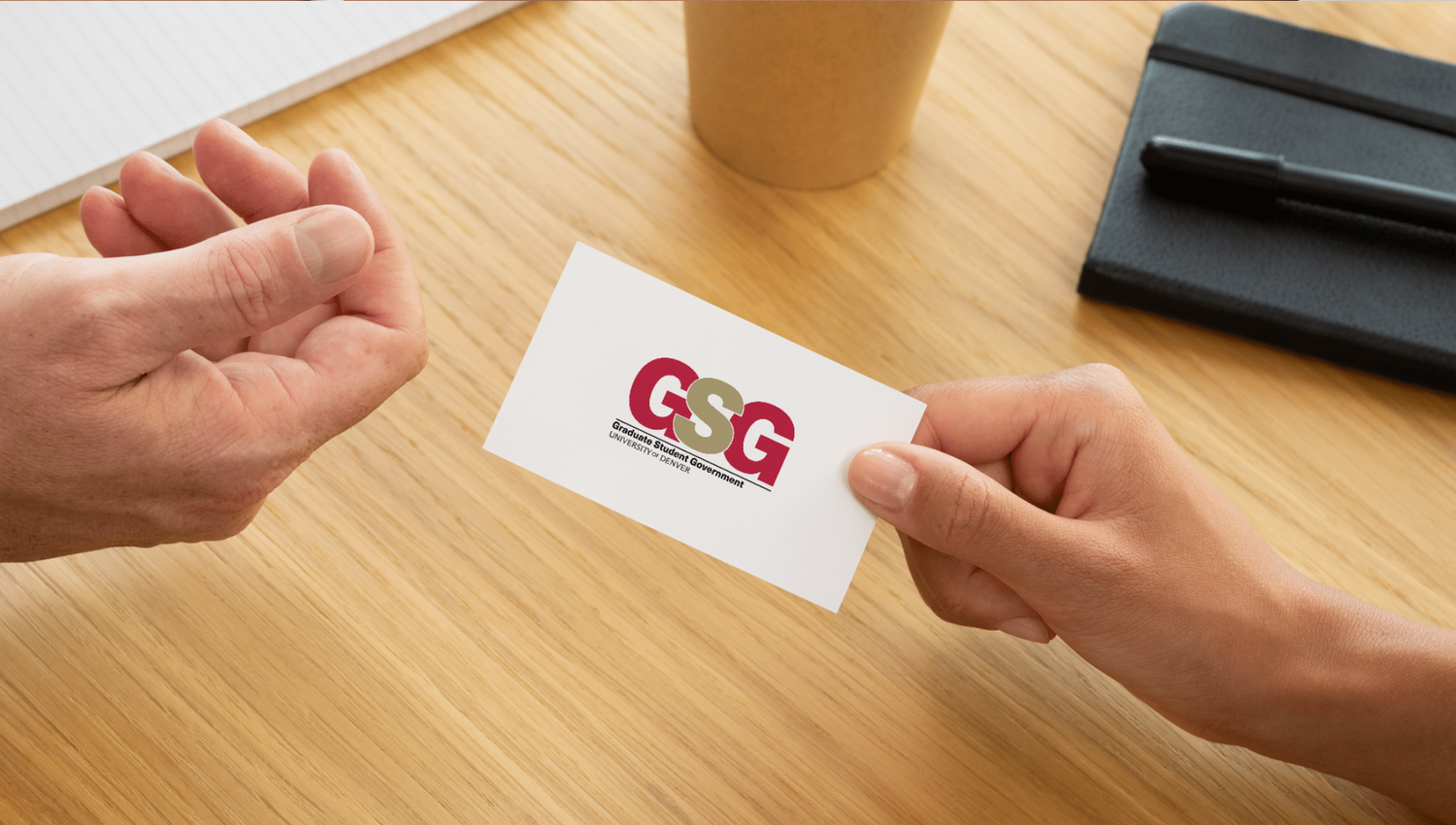 Two hands exchanging a white business card with a colorful GSG logo on a wooden desk surface.