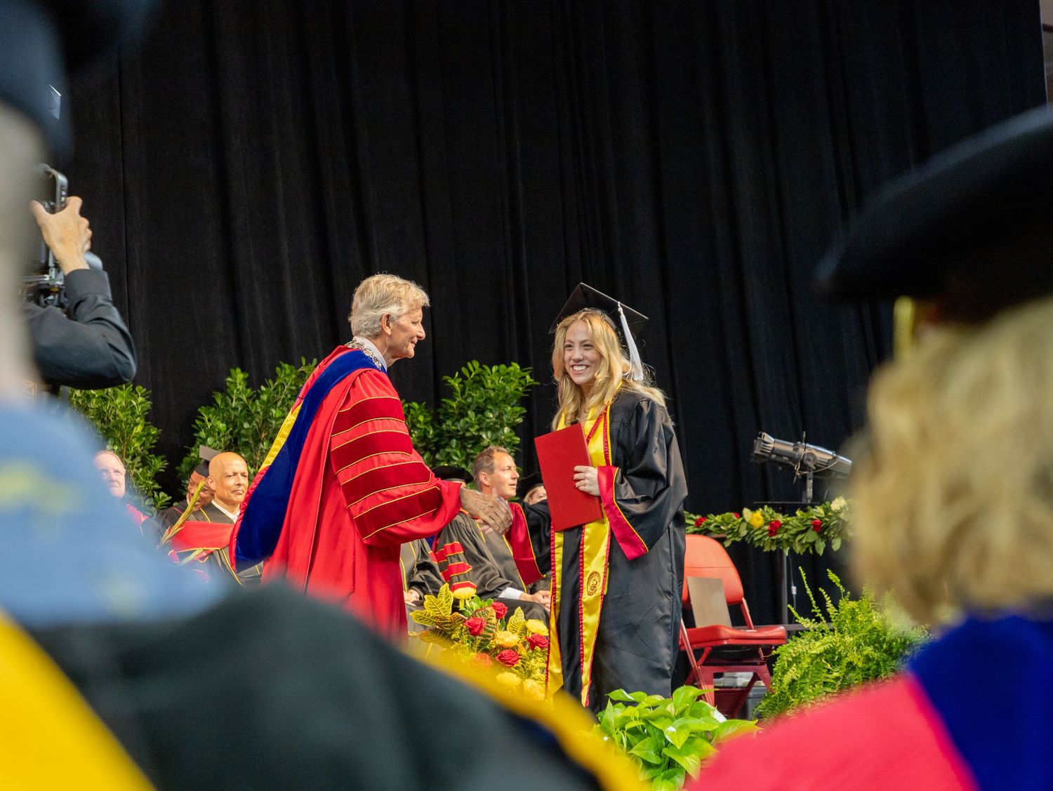 A graduation ceremony with people in red and blue academic regalia gathered outdoors among green foliage.