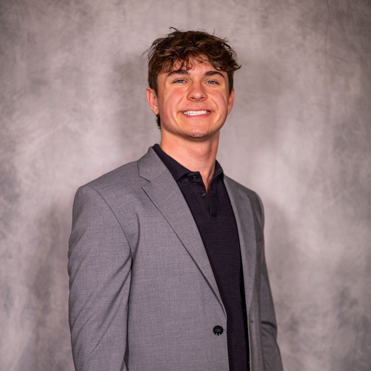 Young man in gray blazer and black shirt smiling confidently against a gray studio background.
