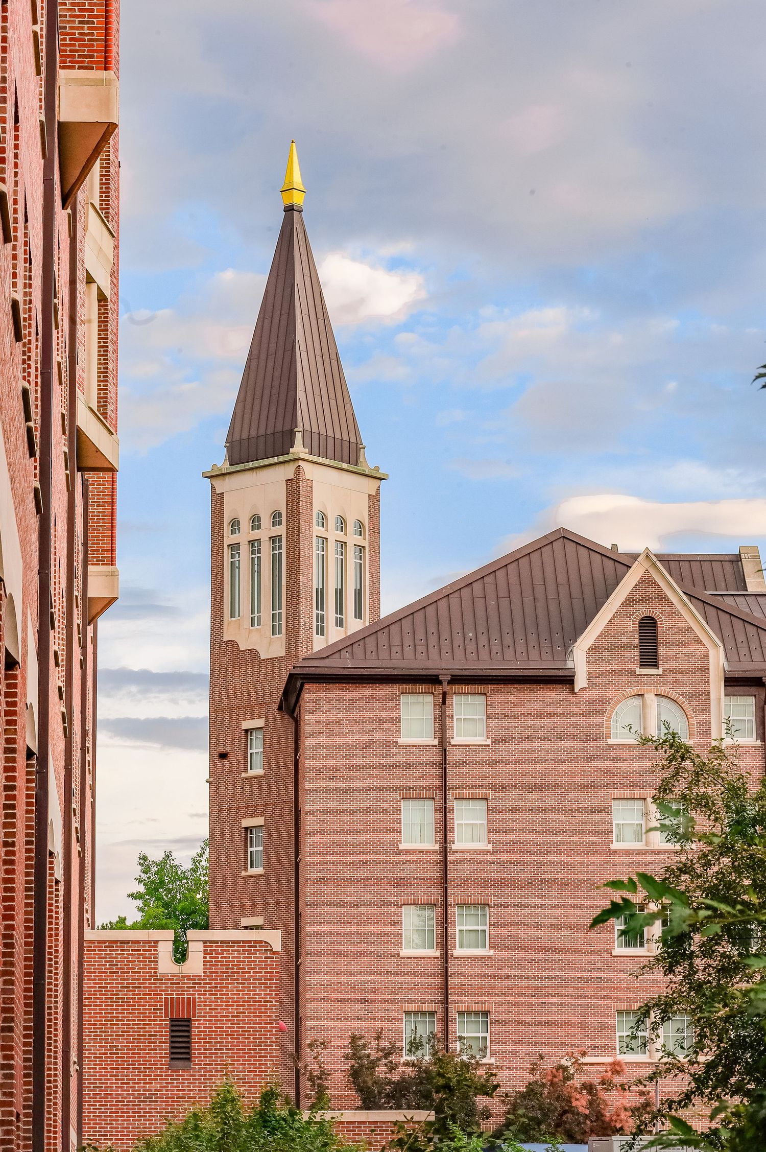 Red brick university buildings with a Gothic-style church tower topped with a gold spire against a partly cloudy sky.