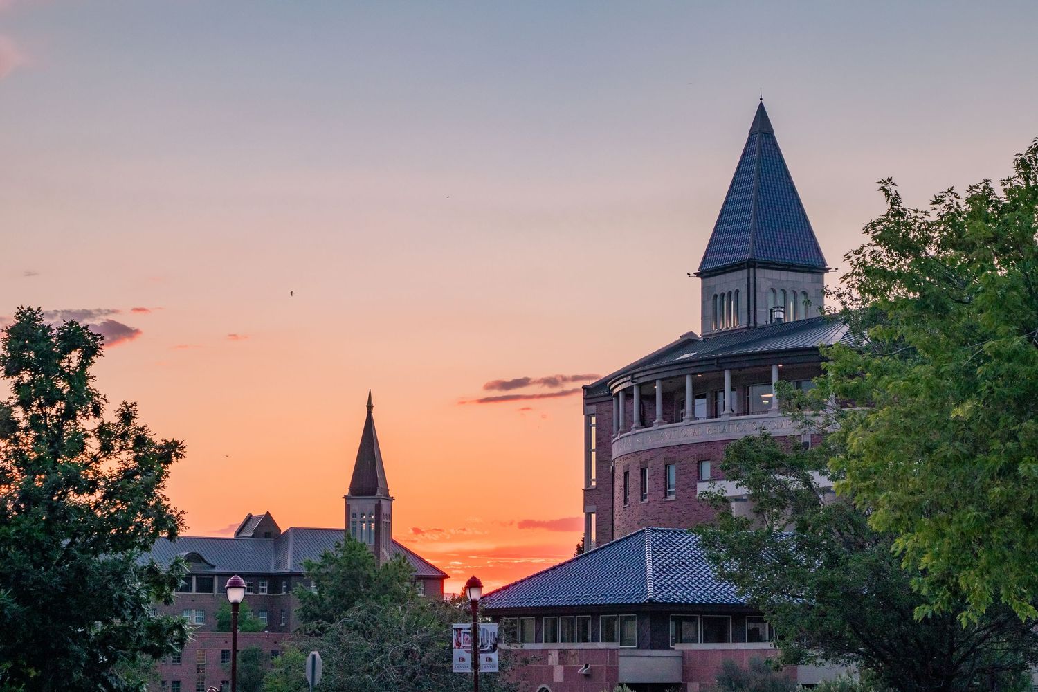 Brick university building with pointed tower silhouetted against vibrant orange and pink sunset sky with trees.